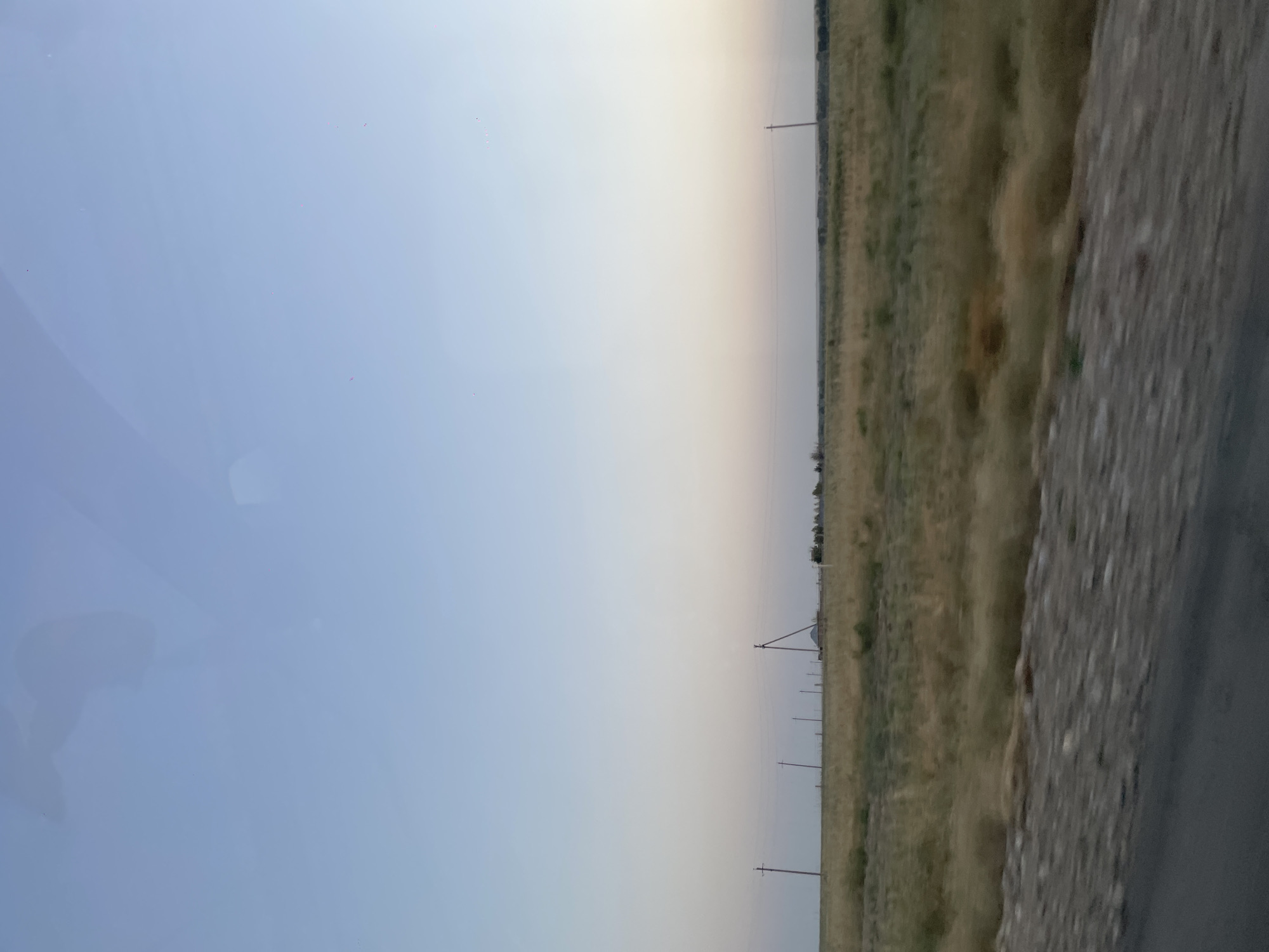 The image captures a vast, open landscape with a clear, pale blue sky transitioning to a soft gradient of pink and white towards the horizon. In the mid-ground, a field of dry, yellowish-brown grass stretches out, dotted with several wooden utility poles with wires strung between them. A low structure, possibly a small building with a pitched roof, is visible behind some of the poles on the right side of the frame. The ground in the foreground appears to be a gravelly roadside next to a dark asphalt surface. The overall scene suggests a rural or semi-arid environment, likely in the city of Toksanbay, Kazakhstan, during either early morning or late evening due to the soft lighting.