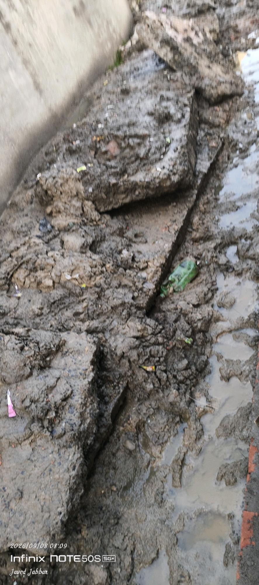 The image shows a muddy, broken-up surface, possibly a construction site or an area affected by recent excavation or damage. Several chunks of concrete or debris are scattered across the scene, with a dark, wet trench cutting through the middle. A green plastic bottle is partially submerged in the mud within the trench. Small pieces of colorful debris, likely trash or litter, are also visible on the surface. The ground appears wet and uneven, with some areas forming small puddles. In the lower left corner, a watermark indicates the date and time "2026/09/08 08:09" and the device "Infinix NOTE50S SG+". The name "Javed Jabbar" is also present, suggesting a potential attribution or photographer. The general environment appears to be an outdoor urban or semi-urban area, possibly in Bahera, India, given the implied context of the watermark.