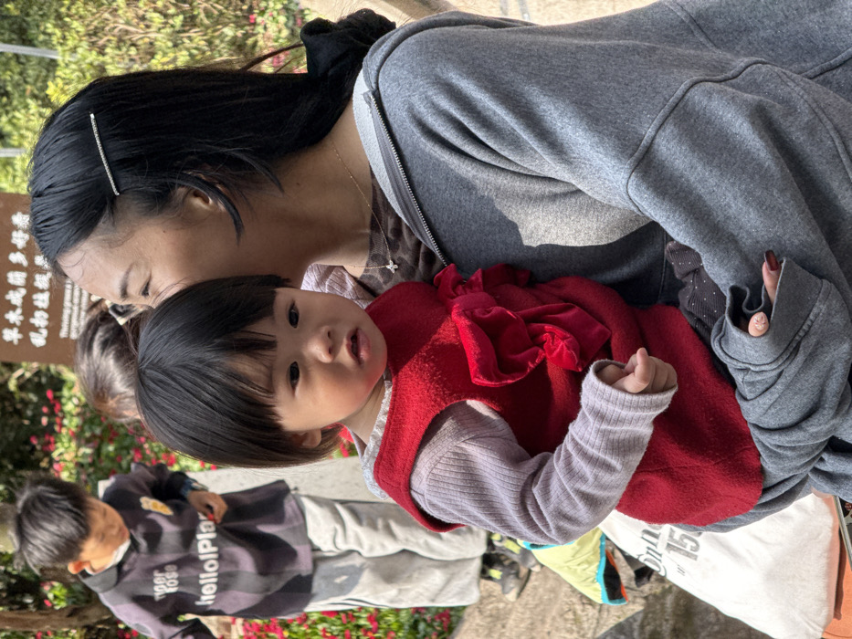 The image captures an outdoor scene in Jiulongpo District, China, featuring three individuals. In the foreground, an adult female with long dark hair, secured by a silver barrette, wears a grey hooded zippered jacket over a brown patterned shirt and a gold chain with a small cross pendant. Her left hand, with dark nail polish, supports a young child, likely a toddler, who has short dark hair and wears a light purple long-sleeved top under a red sleeveless vest with front ruffles. The child looks slightly to the left with a neutral expression. In the mid-ground on the left, a young boy in a dark striped polo shirt with "tiger rose" and "hello! Play" text, along with light grey pants, is seen bending forward, appearing to interact with the ground amidst red flowering plants. A white sign with Chinese characters and partial English text is visible in the upper background. The environment consists of vibrant red and green foliage, a light-colored paved path, and is brightly lit by natural sunlight. A white bag with black text is partially visible on the ground in the lower-left corner.