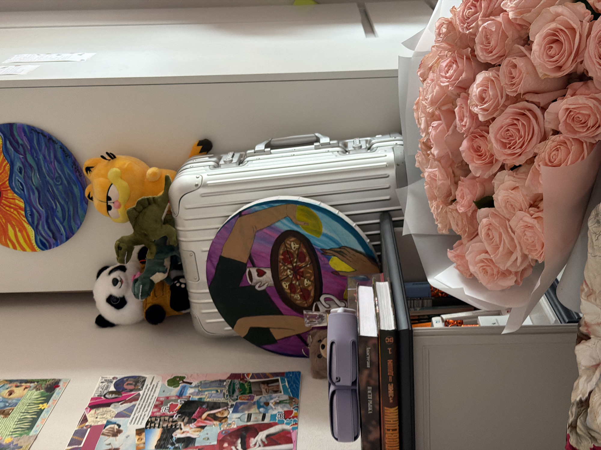 The image captures an indoor arrangement of personal items, decor, and a large bouquet of light pink roses, suggesting a shelf or desk in a personal space.
Dominating the right side is a generous bouquet of delicate, light pink roses, neatly wrapped in white paper. To the left, a silver, hard-shell case, resembling a premium suitcase, is centrally positioned. Leaning against this case are two round art pieces: one features vibrant abstract patterns in blues, oranges, and yellows, while the other depicts a stylized figure holding a pizza and a coffee mug.
Around the case, several plush toys are visible: a yellow Garfield, a green reptilian figure (possibly a dragon), and a black and white panda. On the far right, adjacent to the bouquet and the case, a stack of books stands vertically. One book has a dark brown spine with "DC" at the top and "НИНДЗЯ-ЭЙДЖЕК 1" (NINJA-AGE 1) printed in Cyrillic, which serves as a strong location cue consistent with Moscow, Russia. Another book next to it also appears to have Cyrillic text on its spine. A grey, cylindrical object, possibly a portable speaker or bottle, is positioned near the books.
Below the main display, various printed papers or photographs are scattered, hinting at a collage or collection. The lighting is even, suggesting an indoor setting without specific time-of-day cues. No people are visible, but the collection of items and fresh flowers evoke a personal, cozy atmosphere.