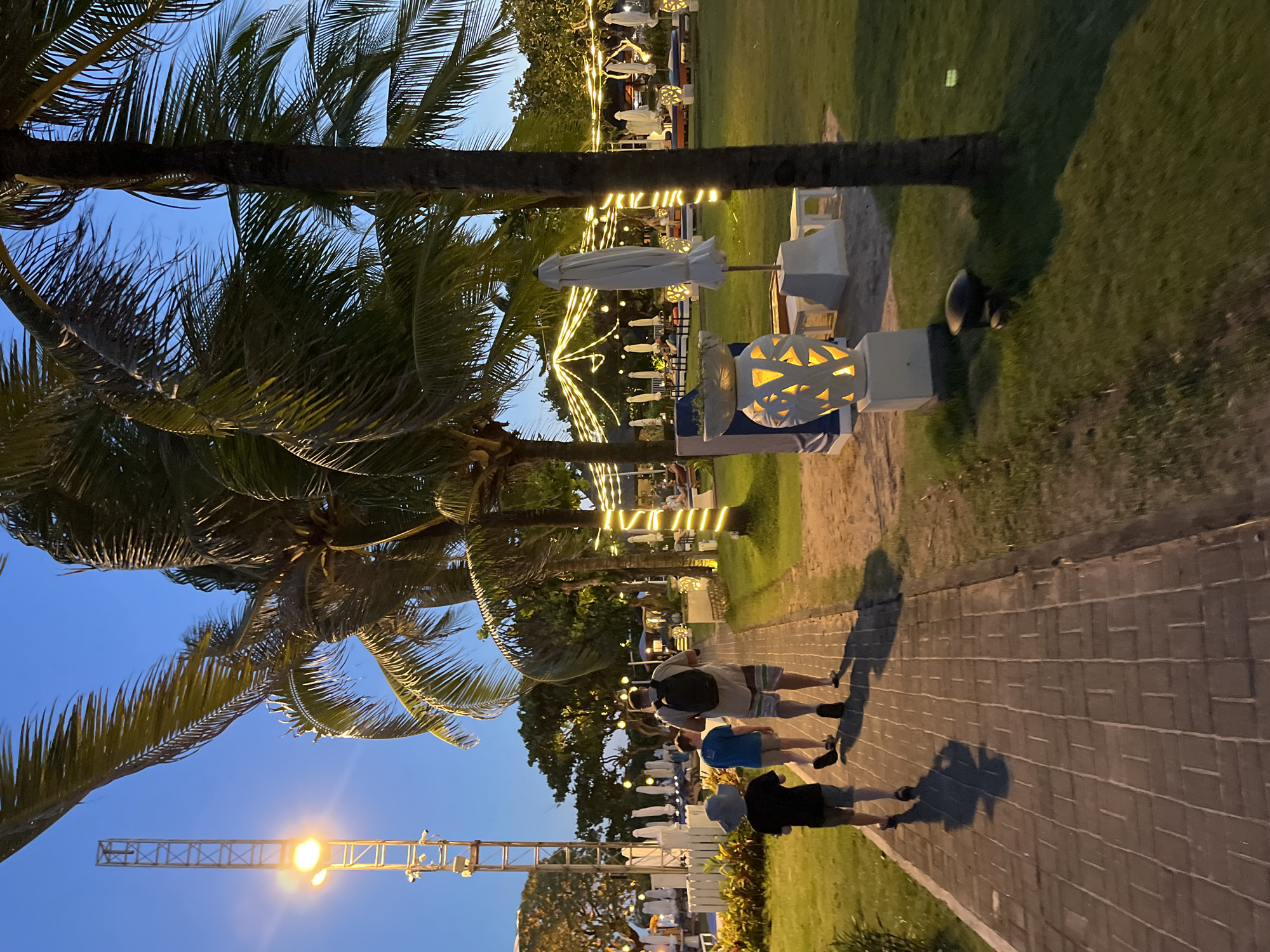 The image shows a street scene in Unknown, Indonesia at dusk. Three people are walking on a brick-paved path. A few people are visible, one with a backpack, wearing shorts. Palm trees line the street with white patio furniture between the walkway and the grass on the right. Patio umbrellas are along the street, with lights strung between the palm trees. A light post is visible on the left side of the image. The sky is light blue.