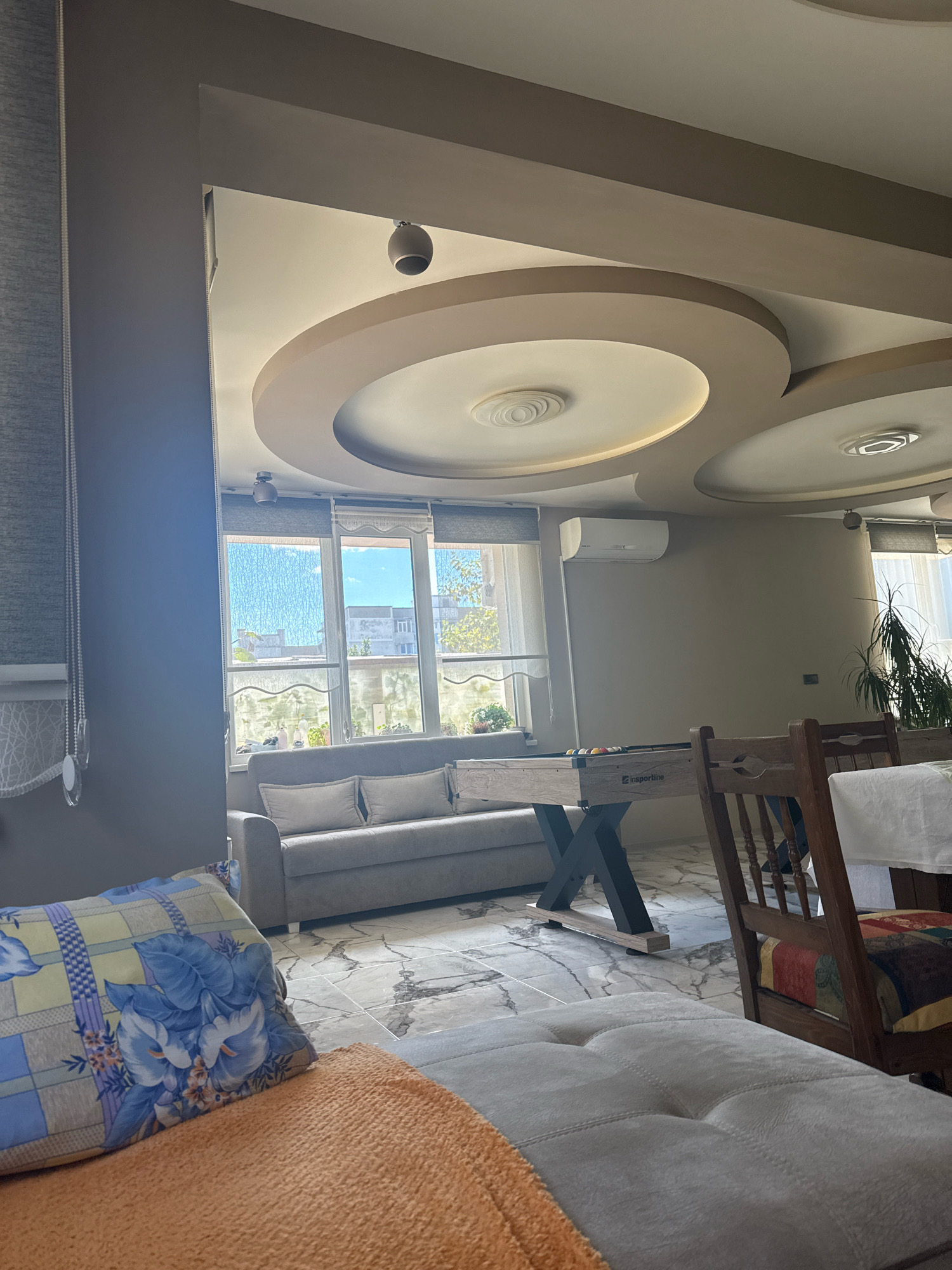 This image shows the interior of a living room or lounge area. The room features a grey sofa, a foosball table, and a dining table with chairs. A patterned pillow and an orange throw blanket are visible in the foreground. The ceiling has a decorative recessed design with lighting fixtures. Large windows look out onto a city scene, suggesting the location is an urban environment. The flooring is a light-colored marble tile. The presence of a foosball table indicates it is a recreational space, possibly within a home, a hotel, or a commercial establishment. The overall impression is one of a comfortable and modern living space.