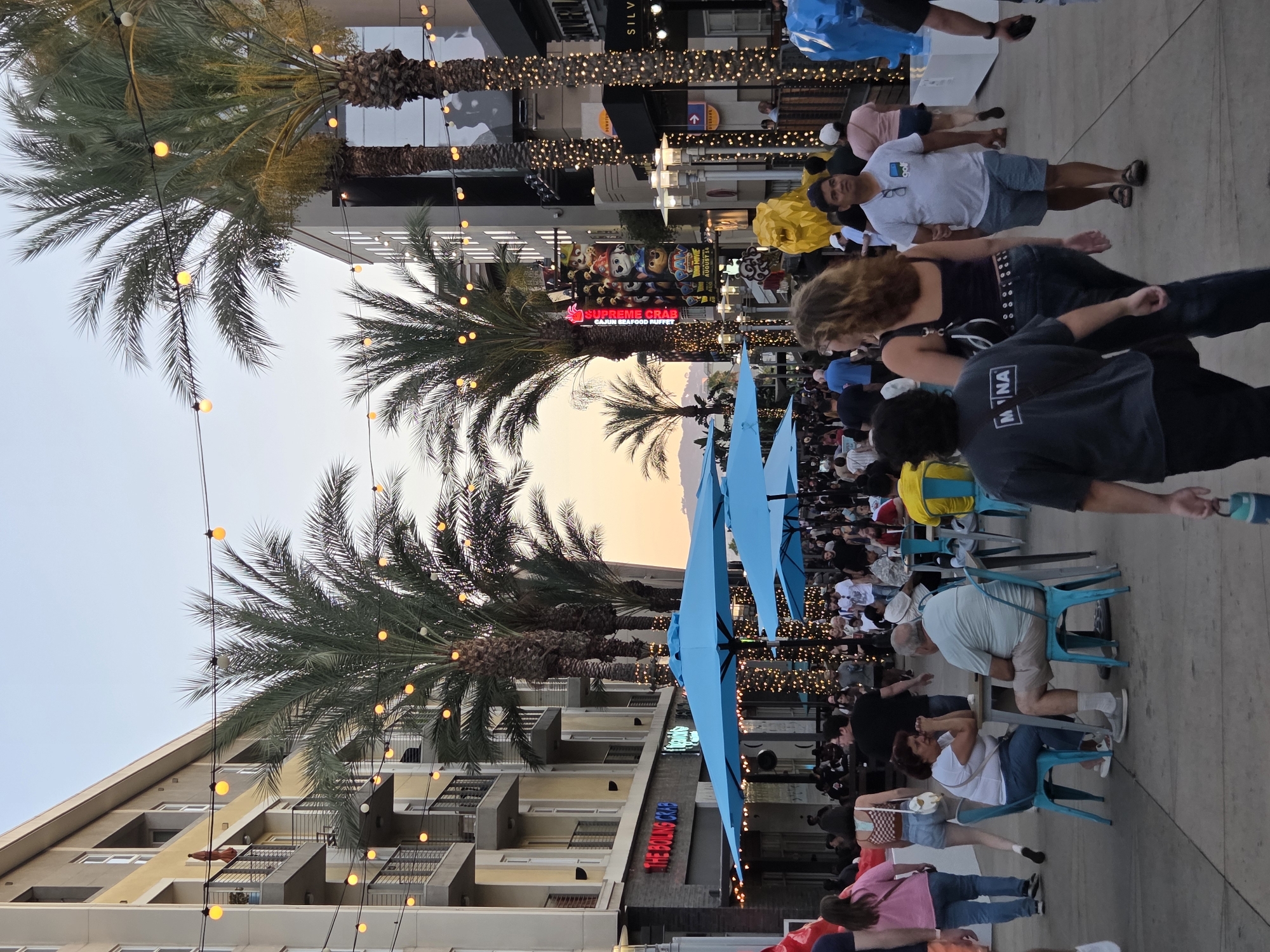 An outdoor pedestrian promenade in Burbank, California, at dusk is depicted. The promenade is lined with multi-story buildings featuring balconies and windows. Palm trees with decorative string lights are interspersed throughout the scene. Numerous people are walking and socializing, with some seated at tables under large blue umbrellas. Businesses are visible, including "THE BOILING CRAB" restaurant. A large vertical billboard displays characters from "Paw Patrol." Decorative elements like a yellow lion statue and blue elephant statue are present. The overall atmosphere suggests a lively evening at an entertainment or dining district.