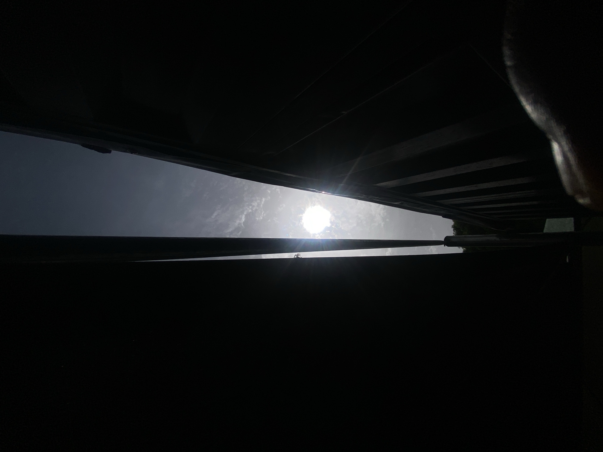The image is taken from a low angle, looking up through a gap in a dark structure. The gap reveals a view of the sky with a bright sun. Parts of what appears to be the structural framework of the building are visible in silhouette in the upper and lower portions of the frame. The location is unclear, but the image indicates that it might be taken in an outdoor setting. No people are visible. It's impossible to verify the specific location within Barden Yana, Nigeria, due to the lack of contextual information.