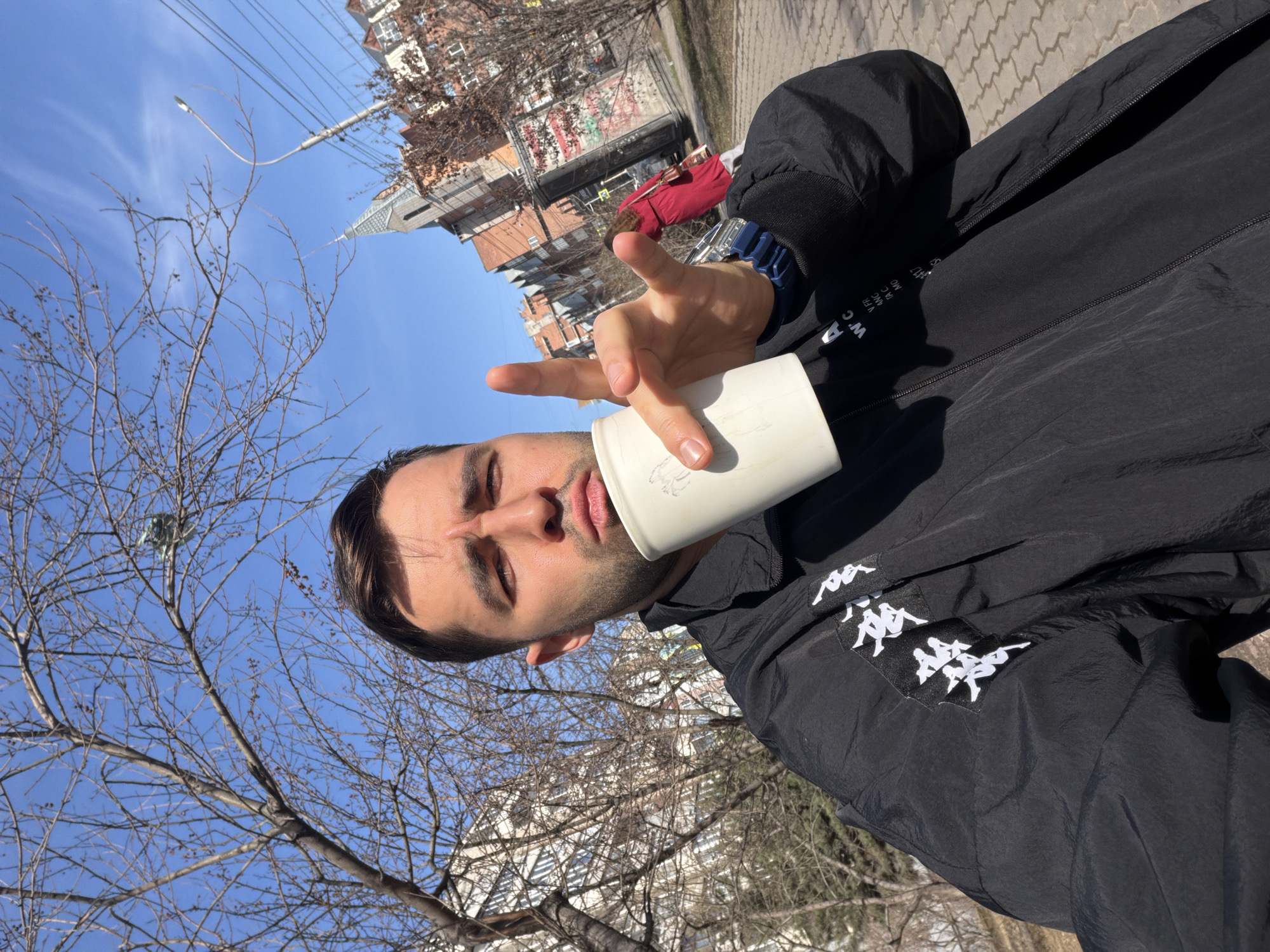 A young man is captured in an outdoor selfie, likely in Krasnoyarsk, Russia, under a bright, clear blue sky. He holds a plain white disposable cup to his lips with his right hand, while his left hand is raised, making a peace sign towards the camera. He wears a black windbreaker-style jacket adorned with multiple white Kappa logos (featuring two figures leaning against each other) on the upper left arm/shoulder. A blue digital wristwatch is visible on his left wrist. Partial white text, including "W.C" and fragmented letters like "FR", "ANC", "MO", is visible near the jacket's main zipper.
The setting is an urban environment, with bare deciduous trees suggesting late winter or early spring. In the background, several residential buildings of various heights are visible, one featuring a distinct pointed roof. Overhead power lines and a streetlight are also present against the clear sky. A paved sidewalk or pathway occupies the lower right of the frame. The man has a calm, neutral expression, contributing to the casual mood of the photograph.
