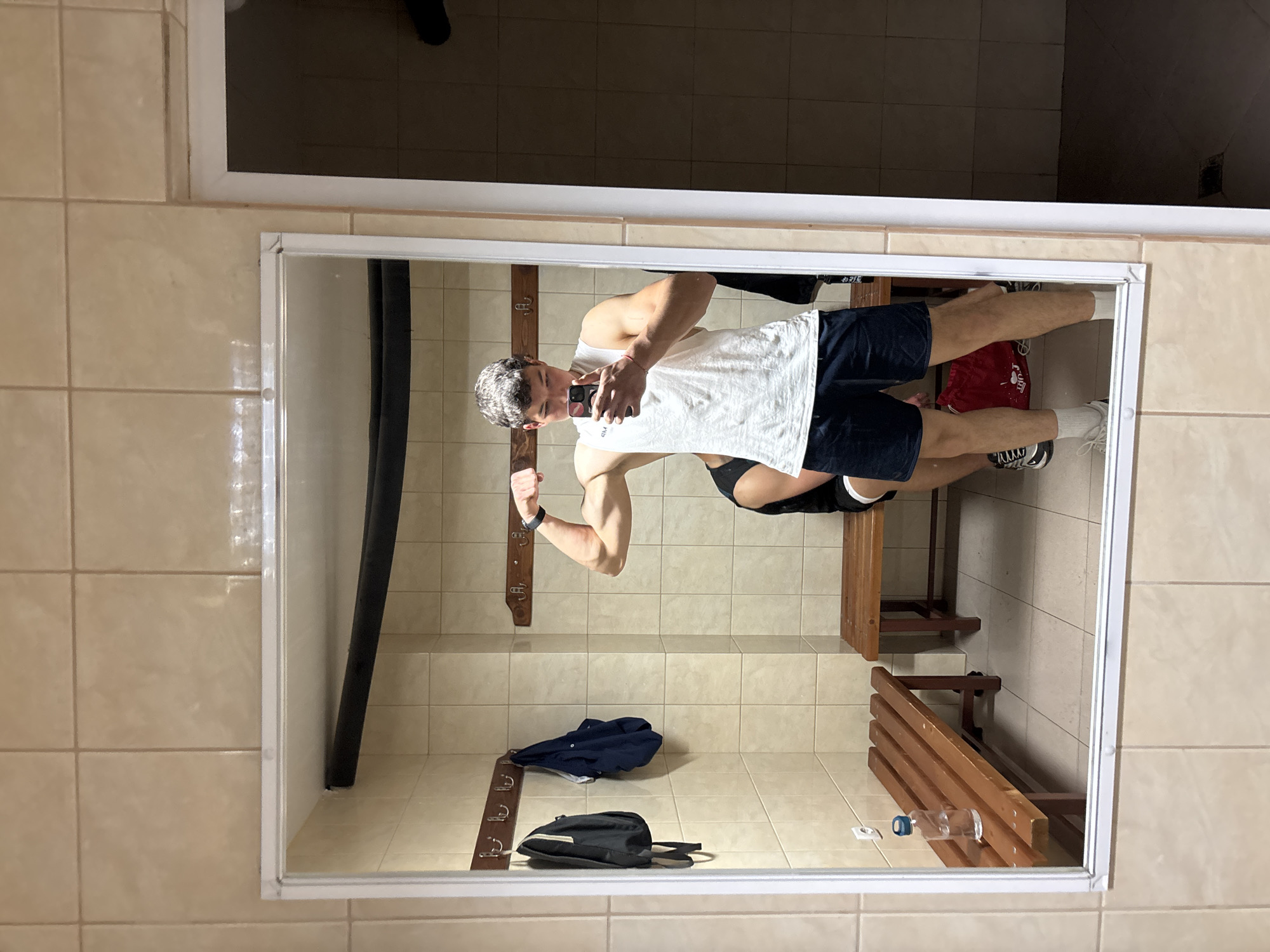 A person is posing in a mirror in a tiled room that appears to be a locker room. The person is wearing a white tank top, navy shorts, and sneakers with white socks. He is flexing his right arm while holding a smartphone in his left hand, presumably taking a selfie. The room contains a bench along one wall, hooks on the wall with some clothes and bags hanging from them. There is also a water bottle on the floor by the bench. The location, Zlatograd, Bulgaria, cannot be confirmed from the image.
