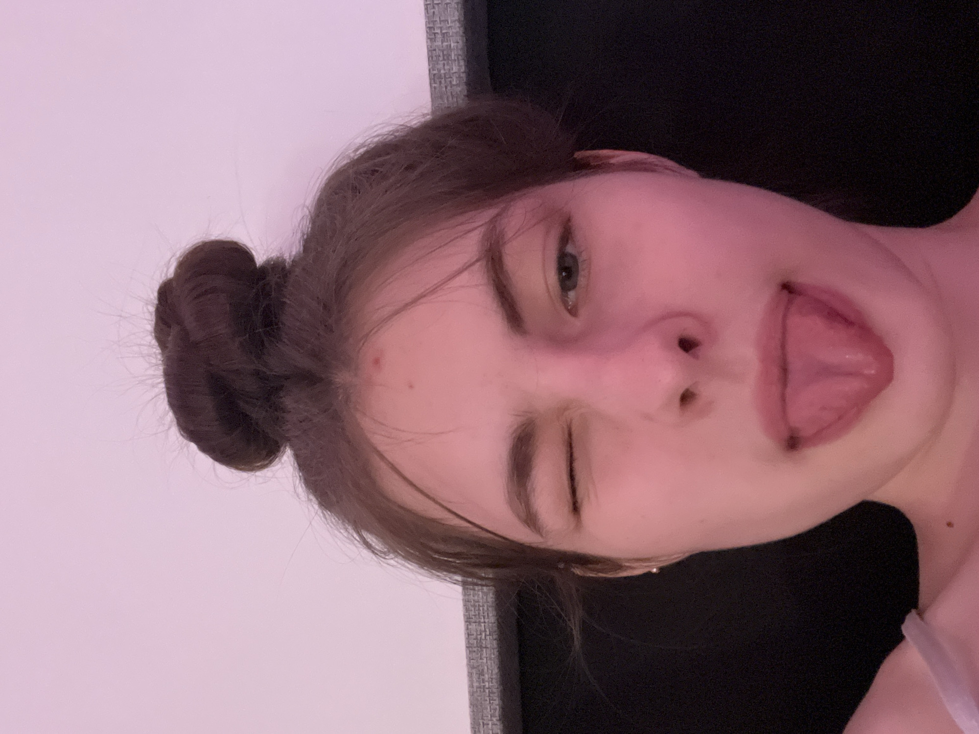 The image shows a fair-skinned young woman with her hair in a bun. She is sticking her tongue out and winking one eye. The background is a combination of a white wall and a dark gray surface. The location is not discernible from the image and could not be identified as Saint Petersburg, Russia.
