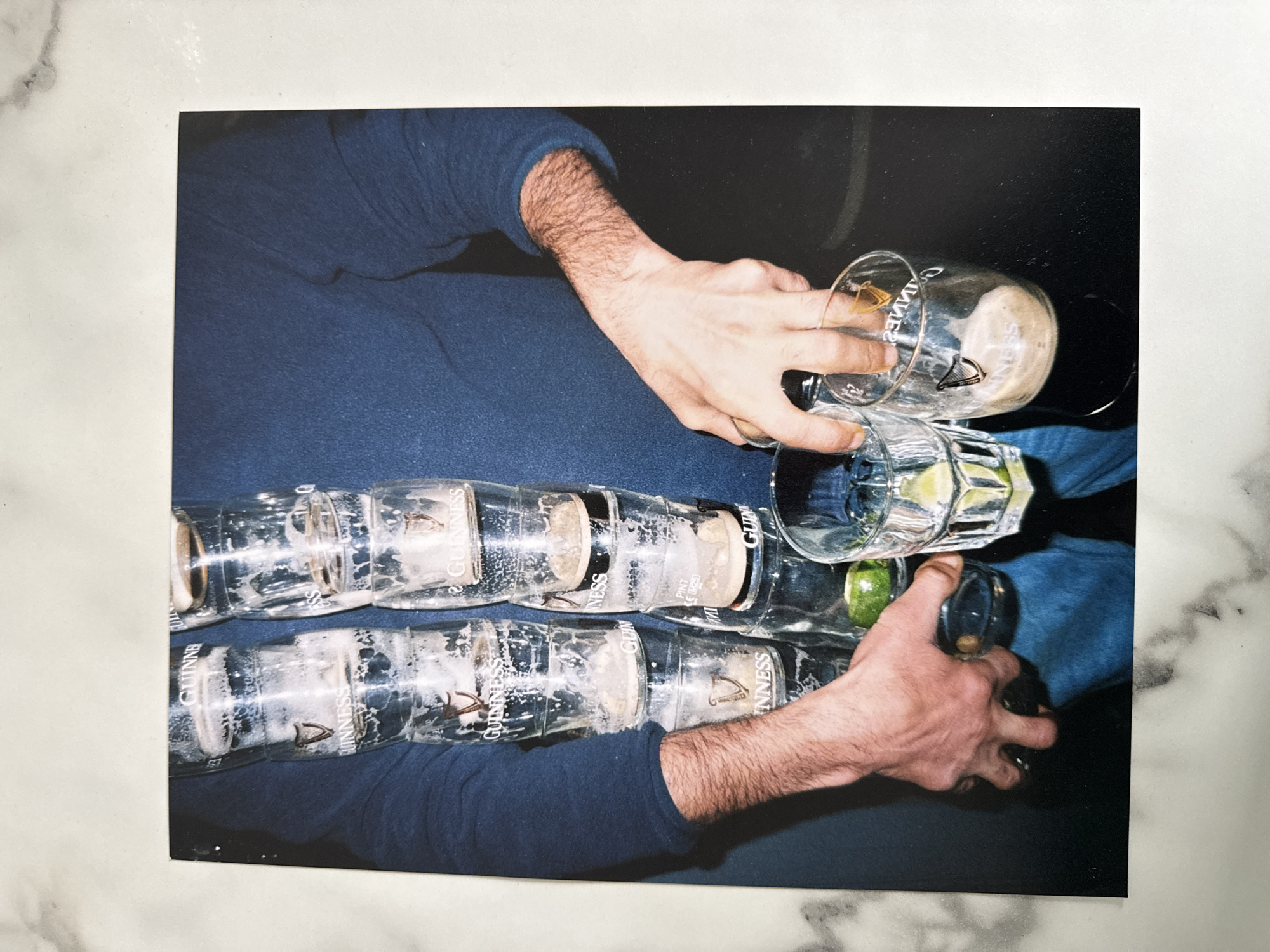 The image shows two rows of Guinness pint glasses. Most of the glasses appear to be empty with remnants of beer and foam. A hand is holding and tilting a full glass of Guinness over a row of glasses that contain lime wedges. The person is wearing a long-sleeved blue shirt. The location in the city of Unknown, Unknown is not evident from the image.