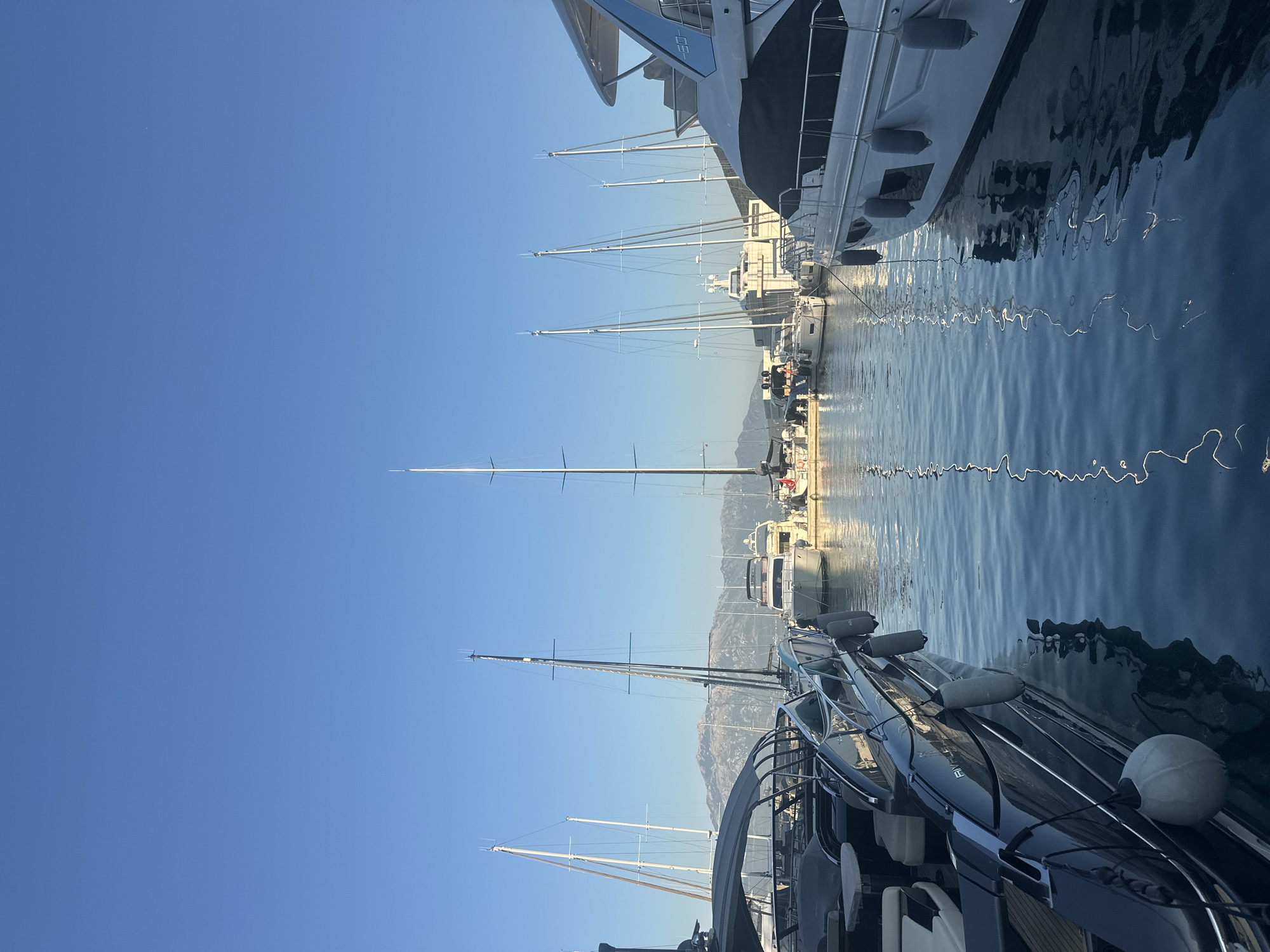 A wide-angle, eye-level shot captures a marina in Herceg Novi, Montenegro, under a clear, bright blue sky. The foreground is dominated by the dark hull of a sleek motor yacht on the left, with several round, gray fenders attached to its side. To the right, a white luxury yacht is docked, its hull also visible with a few fenders. Both yachts are reflected in the calm, dark blue water.
In the middle ground, a line of sailboats with tall, white masts stretches across the frame. Some of these sailboats appear to be moored together. Behind the sailboats, a large, white building or structure is visible, with some people on what appears to be a dock or promenade. Further in the background, more boats and structures are present, and a range of mountains with snow-capped peaks forms a scenic backdrop. The overall impression is one of a tranquil and well-maintained maritime setting.