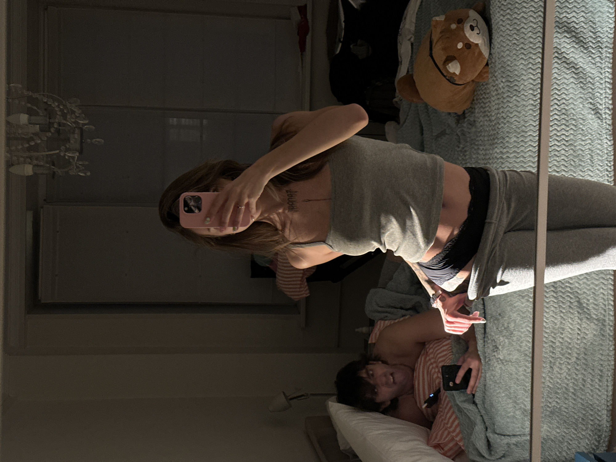 In a dimly lit bedroom, a woman stands facing a mirror, holding a pink smartphone to take a selfie. She is wearing a grey tank top and grey sweatpants with black underwear visible at the waist. A tattoo is visible on her chest. To her left, a man lies in a bed, propped up on pillows, looking at his phone. He is wearing striped pajamas and has an exaggerated expression on his face. The bed is covered with a blue textured blanket. A white pillow is behind the man. A stuffed animal resembling a dog sits on the bed to the right of the woman. The room appears to be an apartment. There is a lamp on a nightstand to the left of the bed. A chandelier hangs from the ceiling, reflected in the mirror. The window behind the bed is covered by blinds.