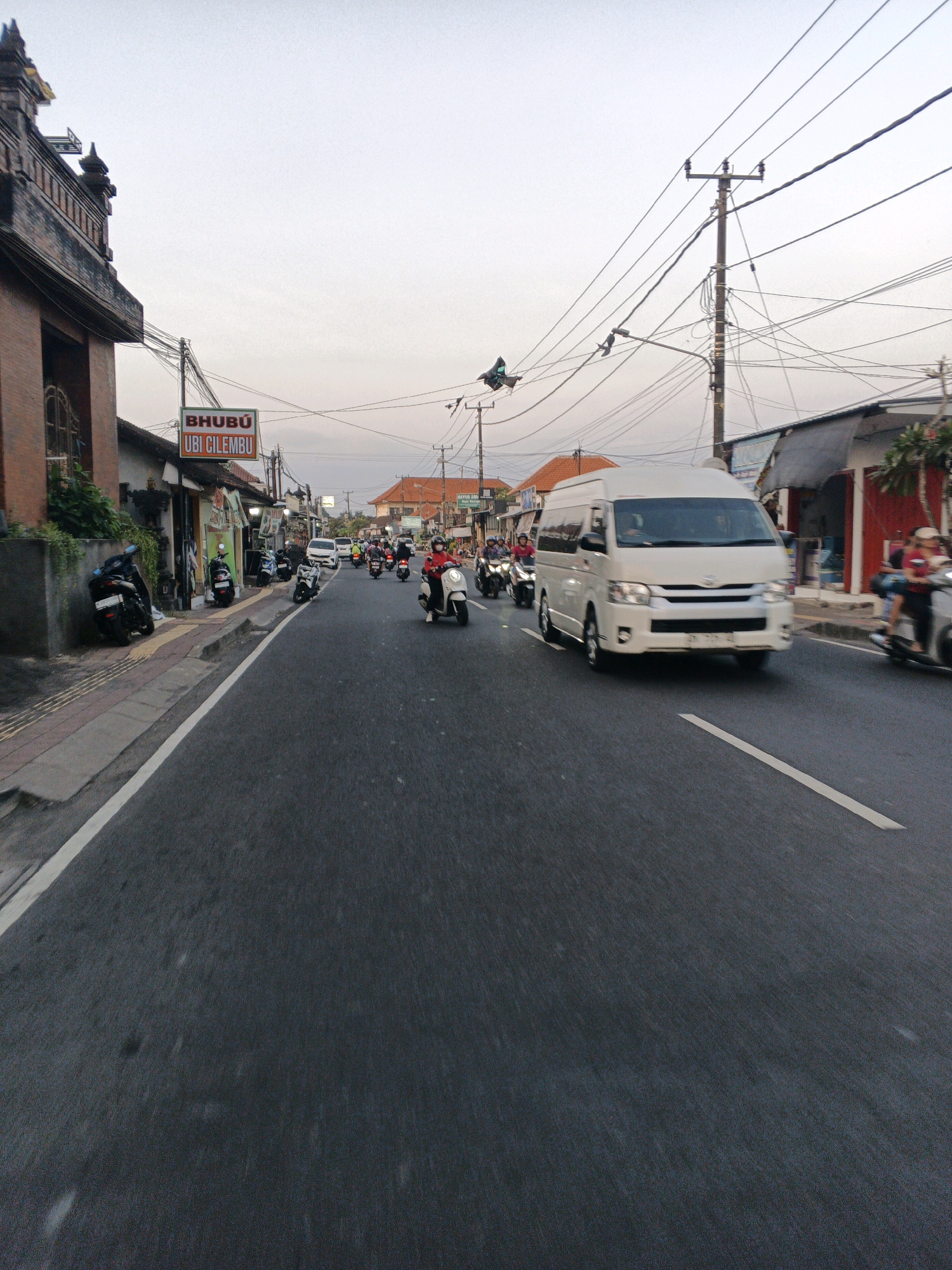 The image captures a street scene in Denpasar, Indonesia, likely during the late afternoon or early evening, given the subdued lighting. The perspective is from a moving vehicle, indicated by the motion blur on the road and surrounding elements.
The street is paved with asphalt and features a white line marking the lane division. On the left side of the road, there is a sidewalk with parked motorcycles. A brick building with ornate architectural details, possibly a temple or traditional Balinese structure, is partially visible on the far left. A sign with red and white lettering, reading "BHUBÚ UBI CILEMBU," is affixed to the building. Further down the street, smaller shops and stalls line the left side.
The right side of the street is also lined with buildings, some with red facades and tiled roofs. Overhead, a complex network of electrical wires is supported by wooden poles, a common sight in urban Indonesian areas.
Several vehicles are present on the road. A white van is prominently positioned in the right lane, appearing to be in motion. Numerous motorcycles and scooters, many with riders wearing helmets and some in bright clothing, are also navigating the street, indicating a busy thoroughfare. One scooter rider in a red top is particularly noticeable in the right lane.
The sky is overcast with a pale, diffused light. A green kite is visible suspended from one of the electrical wires in the distance. The overall atmosphere suggests a typical day of traffic and commerce in an Indonesian city.