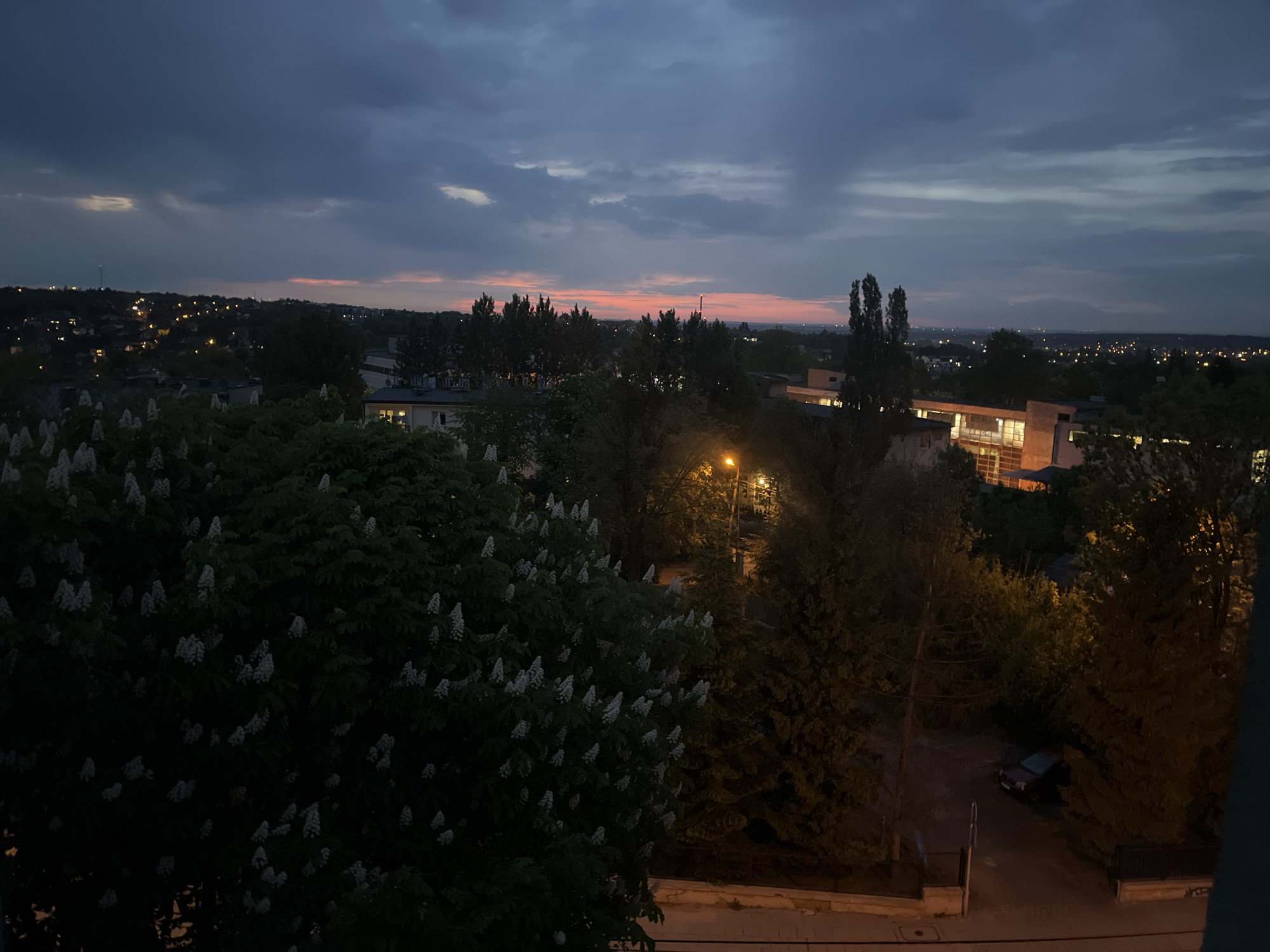 The image shows an evening urban landscape in an unknown city. In the foreground, a large tree with white blossoms fills the left side of the frame. Streetlights illuminate trees and buildings in the middle ground, casting warm, yellow light. A car is parked near the edge of the scene. In the background, buildings and structures are visible, with scattered lights suggesting residential areas. The sky is a mix of dark blues and purples, with a hint of pink on the horizon, indicating sunset or twilight. The overall atmosphere is quiet and calm.