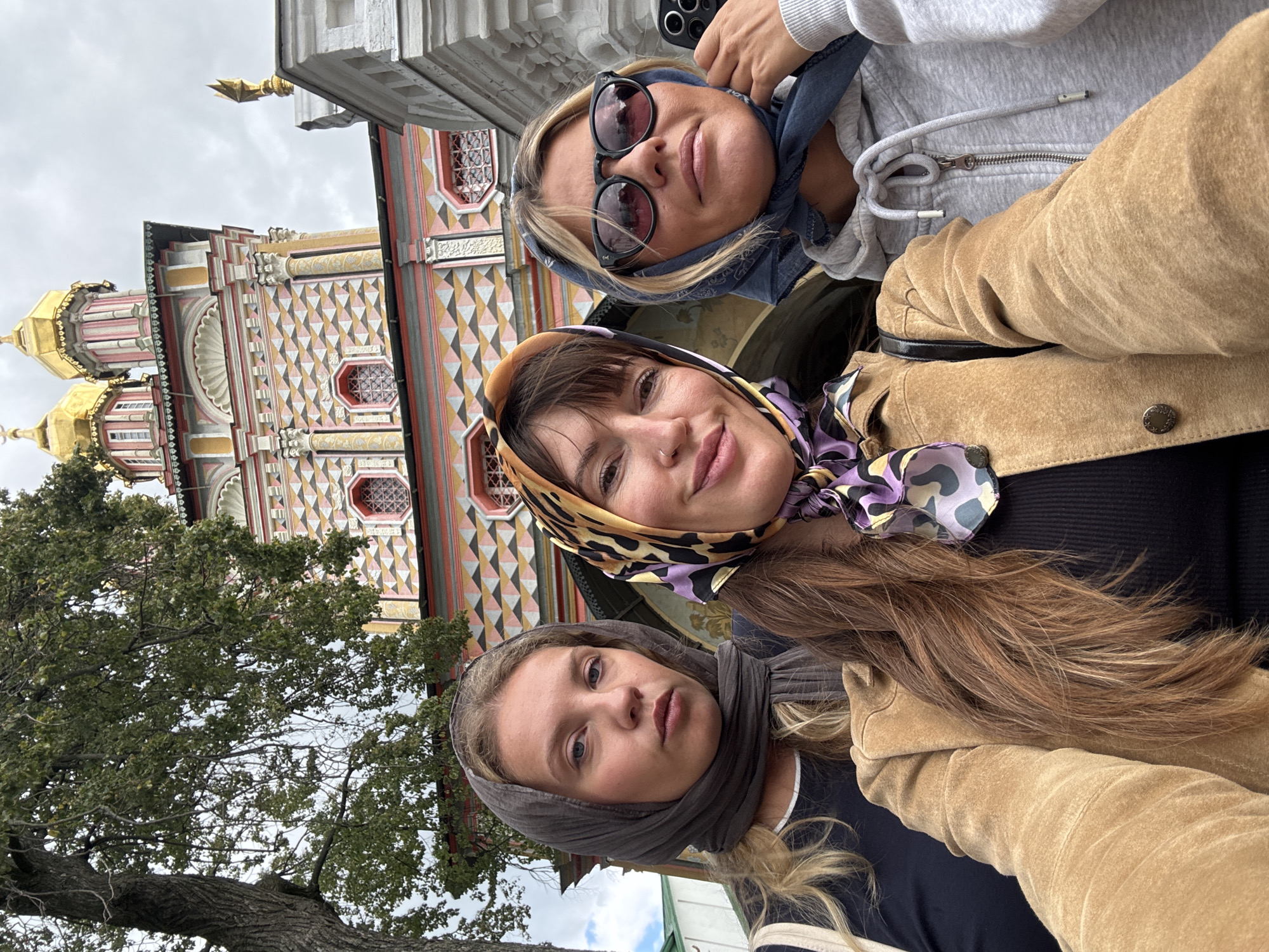 Three women are taking a selfie in front of a brightly colored, ornate building with golden domes. The woman on the left is wearing a dark grey headscarf and a black shirt. The woman in the center is wearing a leopard and purple patterned headscarf, a black ribbed top, and a tan suede jacket. The woman on the right is wearing a blue headscarf, sunglasses, and a grey hooded sweatshirt. The building behind them appears to be a church or monastery, with intricate geometric patterns and arched windows. The overall scene suggests a visit to a cultural or religious site in Sergiyev Posad, Russia.