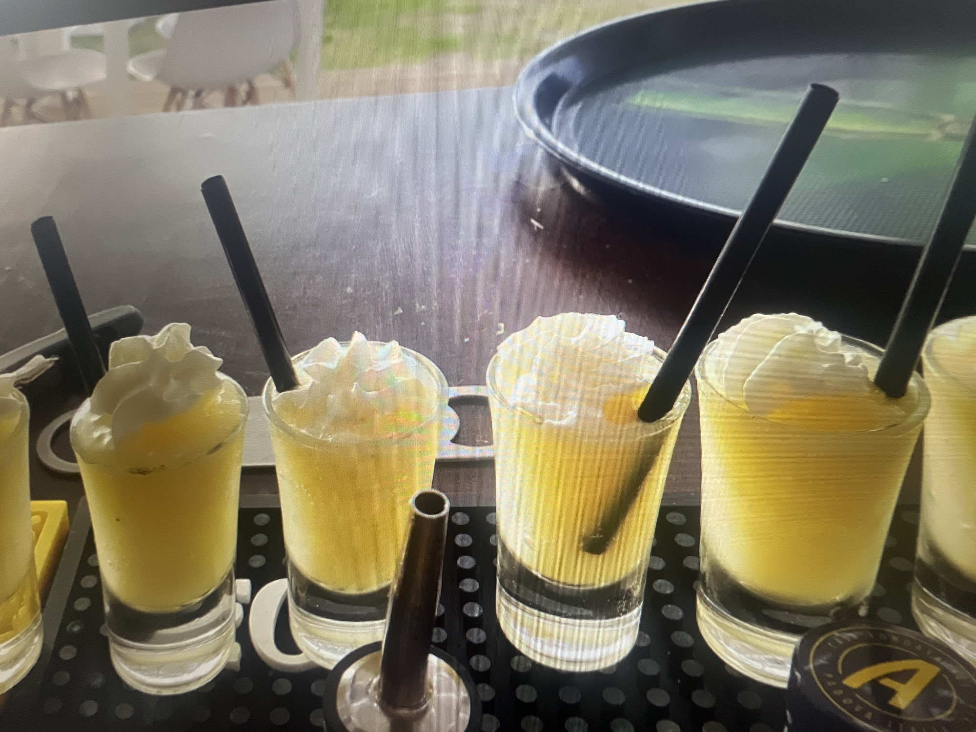 The image depicts a bar scene featuring several shot glasses filled with a yellow, possibly citrus-based, beverage topped with whipped cream and garnished with a black straw. The shot glasses are arranged on a black bar mat, which has a pattern of small circles. A metal spout or stopper is positioned between the glasses. In the background, a dark-colored serving tray and some blurred outdoor scenery are visible. The details in the image are not sufficient to identify a specific location in Sofia, Bulgaria.