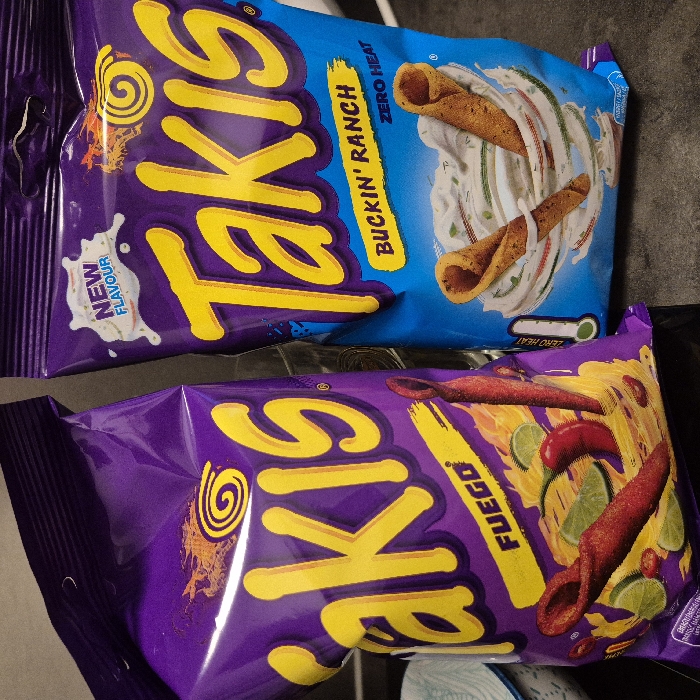 The image displays two upright, packaged bags of "Takis" brand rolled tortilla chips on a dark surface in Strasbourg, France. The left bag, predominantly purple, features the "Fuego" flavor with yellow lettering, depicting red chili peppers, lime wedges, and red rolled chips amidst a fiery design. The right bag, primarily blue, is labeled "Buckin' Ranch" with yellow lettering and a "NEW FLAVOUR" indicator, showing tan rolled chips enveloped in a white ranch dressing swirl, accompanied by a "ZERO HEAT" thermometer icon. A small, light-colored patterned bowl is partially visible to the far left behind the purple bag. The background is dark and out of focus.