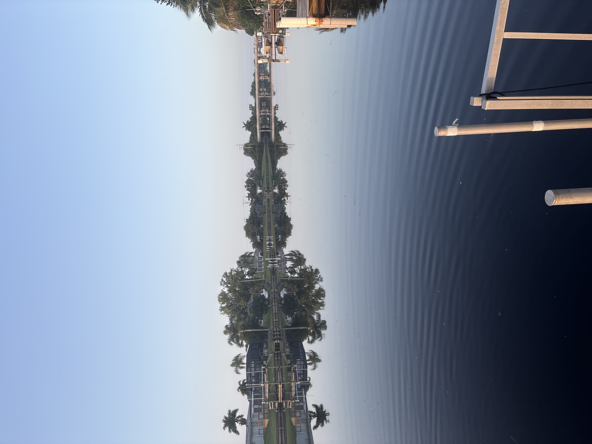 This image depicts a tranquil canal scene in North Port, Florida, USA, likely during the early morning or late afternoon given the soft lighting and clear sky. The water is calm, reflecting the houses, palm trees, and vegetation lining the opposite bank. A bridge spans the canal in the distance. The foreground shows a portion of a dock or pier with metal railings and support posts. The environment is residential and suggests a waterfront community.
