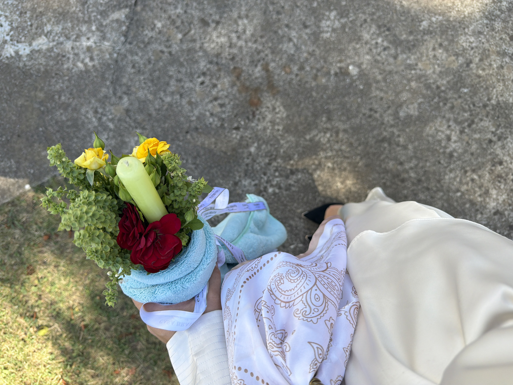 A person, viewed from above, is holding a bouquet of red and yellow roses, green foliage, and a light green cylindrical object. The bouquet is wrapped in a light blue towel and tied with a white ribbon. The person is wearing a cream-colored outfit, a white patterned scarf, and black shoes. They are standing on a textured gray surface, possibly a sidewalk or pavement, with a patch of green grass and dry leaves visible in the background. The scene appears to be outdoors, with sunlight casting shadows. There are no visible landmarks that specifically indicate the city of Zăluceni, Moldova.