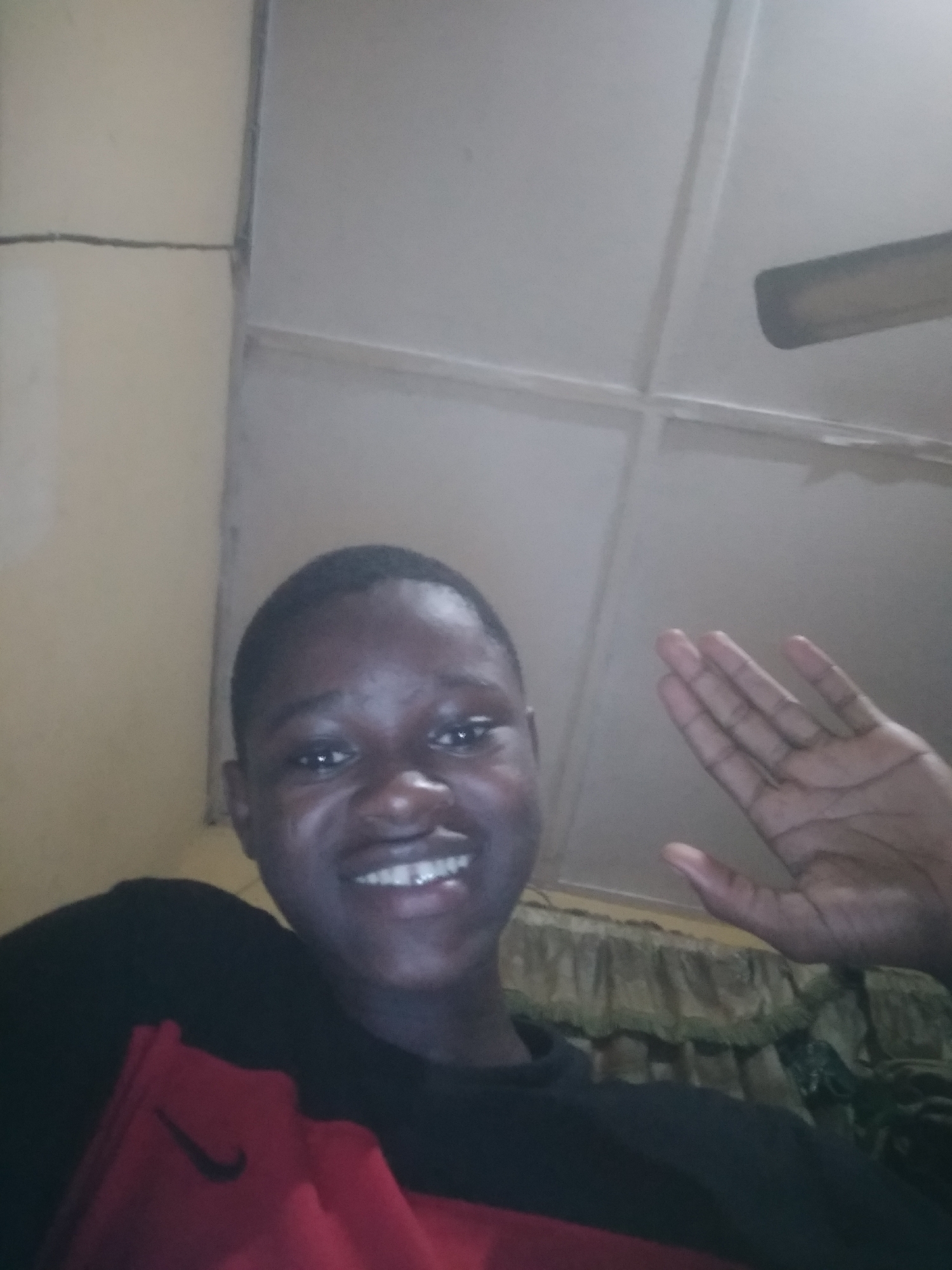 A young person with short hair is smiling and waving. The person is wearing a black and red Nike shirt. The background consists of a ceiling with a square panel design and a partial view of what appears to be a curtain. The location is presumably indoors but the city is not possible to determine based on the provided information.