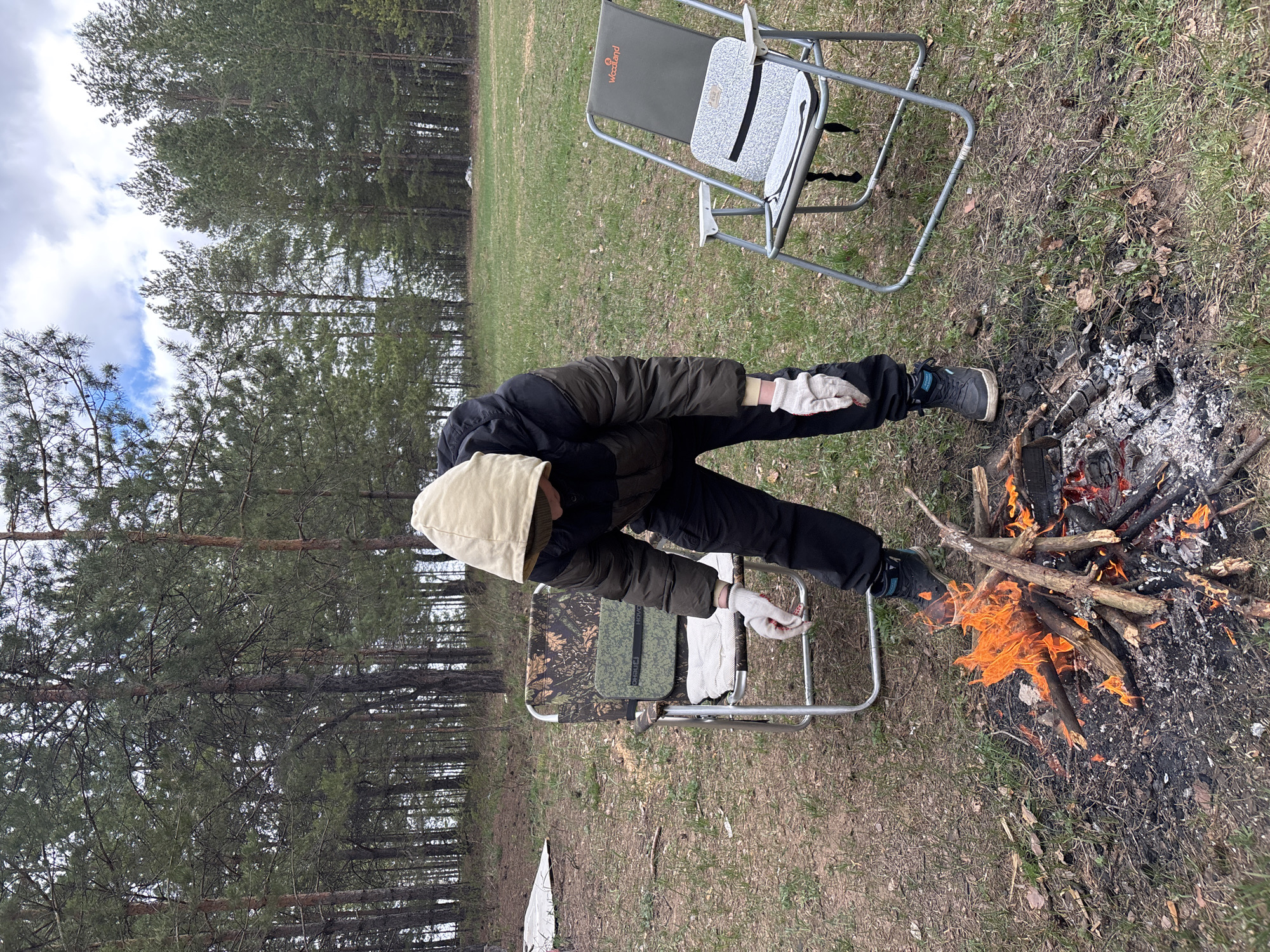 A person is tending to a campfire in what appears to be a wooded area, possibly on the outskirts of a city, Vekoshinka, Russia, based on the prompt. The individual, wearing a dark jacket, black pants, and light-colored gloves and a hooded hat, is bending down near the fire. The fire is actively burning with flames. Two folding chairs are nearby, one slightly behind and to the right of the person and another closer to the person and the fire. The ground is covered with grass, dry leaves, and burnt wood. Trees are visible in the background.