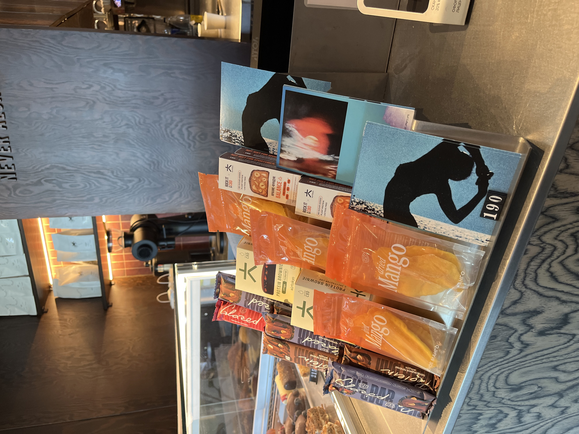 The image shows a counter display in a cafe. On the counter, there are several rows of packaged goods, including what appear to be protein bars and dried mangoes. Behind these items, there are some printed cards with abstract art and silhouettes. A coffee machine is visible in the background. The cafe has a modern design with wooden accents and soft lighting. There are no people visible in the image.