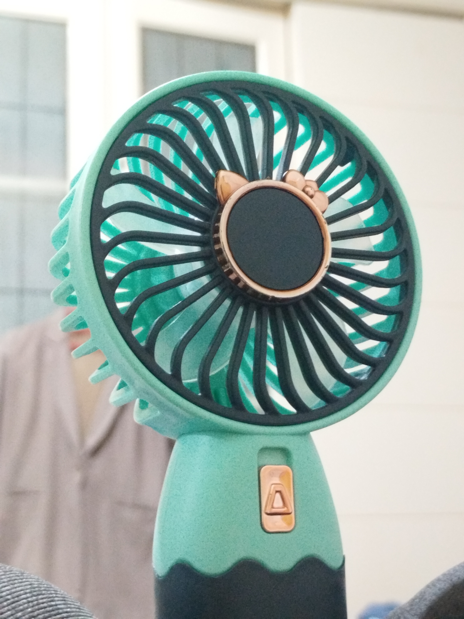 A close-up shot shows a small, handheld fan with a light green plastic frame and a black central circular area. The fan's blades are visible through the safety grill. The handle is a darker shade of green at the bottom, blending into black. The location and the city of Lhokseumawe, Indonesia, are not discernible from the visual information in the image. No people are clearly visible. The environment seems to be indoors, suggested by the blurry background that shows a window and a person in a light-colored shirt.
