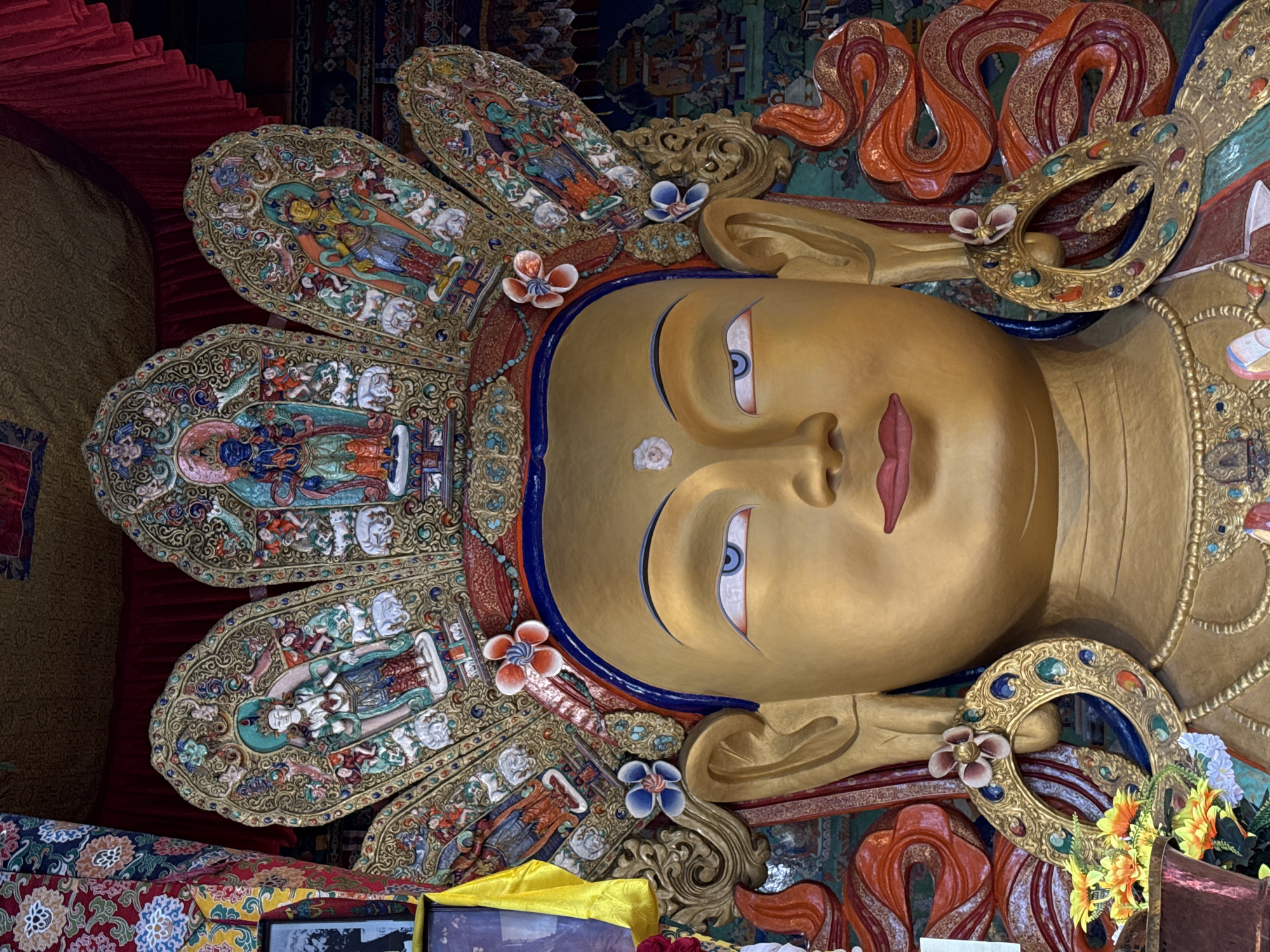 The image shows a close-up of a large, golden Buddha statue within the Thiksey Monastery in Leh, Ladakh, India. The statue is adorned with an elaborate, multi-tiered crown featuring intricate carvings and depictions of various figures. The Buddha has serene blue eyes, a urna on its forehead, and full red lips. Ornate gold earrings with colorful gemstones hang from its ears. The background is a richly decorated tapestry with vibrant patterns and religious imagery. The statue is positioned in what appears to be the main assembly hall of the monastery.