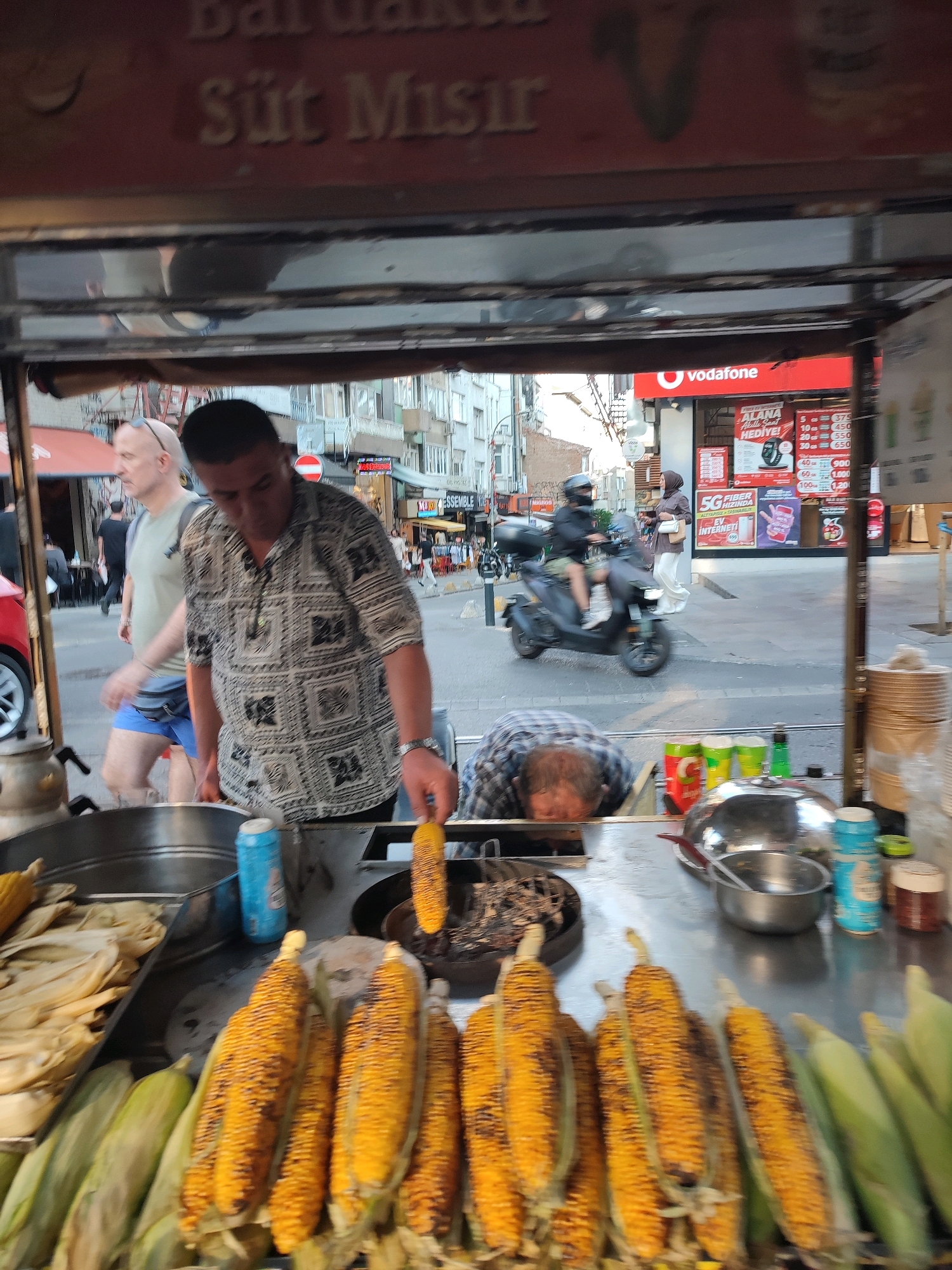 The image captures a street scene in Kadıköy, Turkey, focusing on a vendor selling corn from a cart. In the foreground, several ears of grilled corn are displayed prominently, some still in their husks. A vendor, wearing a patterned shirt, is seen holding a cob of corn, possibly preparing it for a customer or placing it on a grill. In the background, other people are visible, including a man walking by in a light-colored t-shirt and blue shorts, and a person on a scooter passing by. The setting appears to be a busy urban environment with buildings and shops lining the street. A Vodafone advertisement is visible on a building in the background, further indicating a commercial area. The overall impression is of a typical street food vendor operation in a bustling Turkish city.