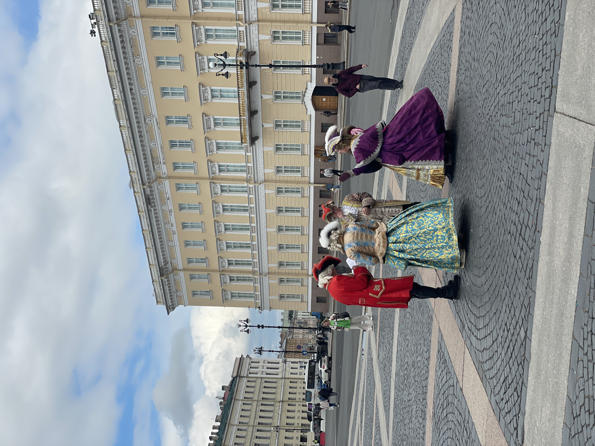 Three individuals dressed in elaborate historical costumes are standing on a cobblestone square. The man on the left wears a red coat with gold trim and a tall black hat. The person next to him wears a light blue and gold gown with fur trim. The person on the right wears a purple dress and a white hat. The background is dominated by a tall, pale yellow building with multiple windows and a decorative facade. There are street lamps and other buildings visible in the distance, along with cars and pedestrian traffic. The sky above is blue with scattered clouds. This photograph appears to be taken in Palace Square in Saint Petersburg, Russia, a popular tourist destination.