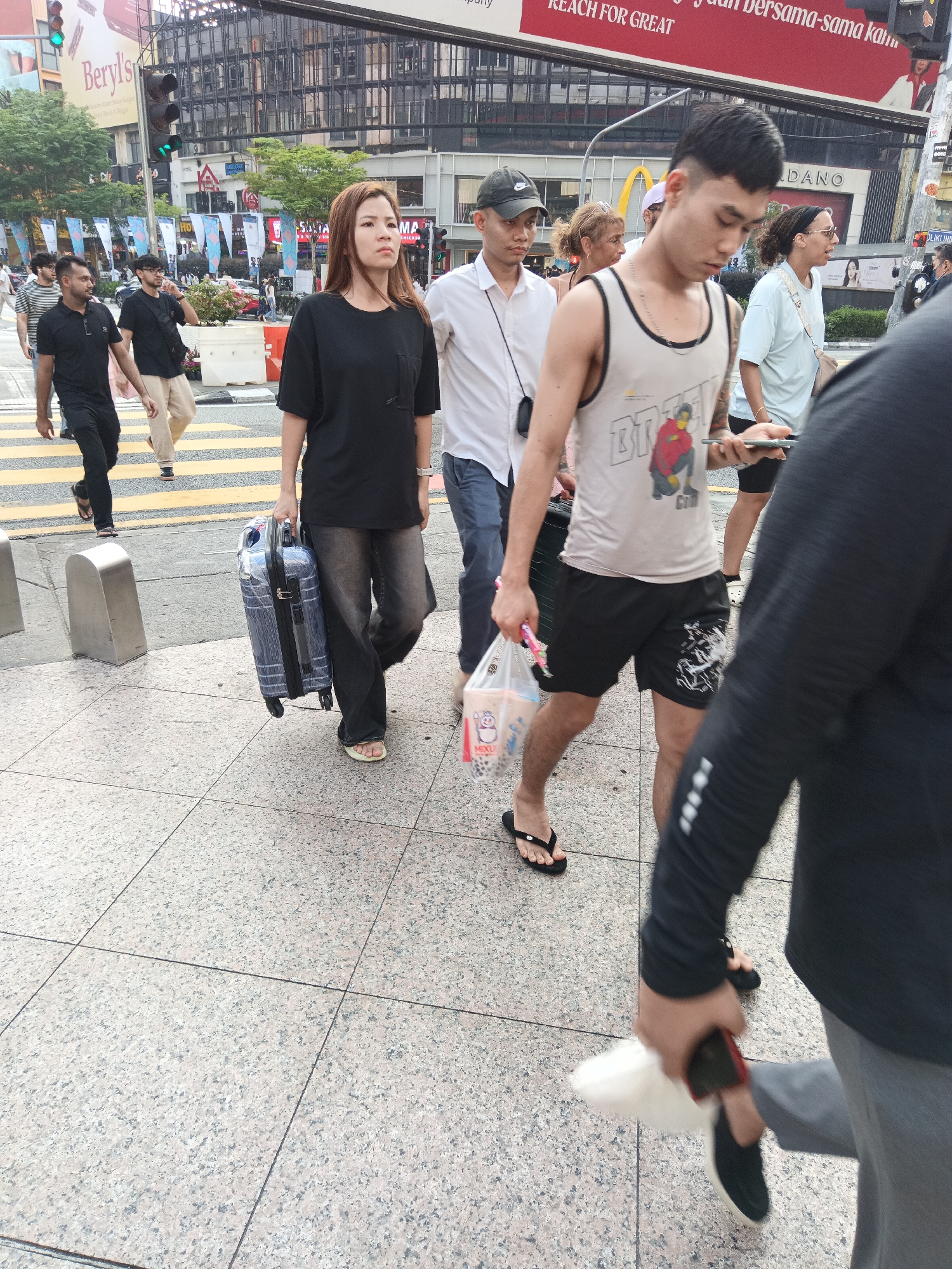 A group of people are walking on a pedestrian crossing in Kuala Lumpur, Malaysia, with a street and buildings in the background. The individuals are diverse in gender and ethnicity. A woman in a black t-shirt and dark pants is pulling a blue suitcase. Beside her, a man in a white shirt and dark pants is looking at his phone. To the right, a young man in a white tank top and black shorts is walking with a bag of drinks. Several other individuals are also visible, some walking in the same direction as the main group. The environment includes traffic lights, signage, and a busy urban streetscape. The pedestrian crossing is marked with yellow lines. The weather appears to be overcast or it is late in the day as there are no harsh shadows.