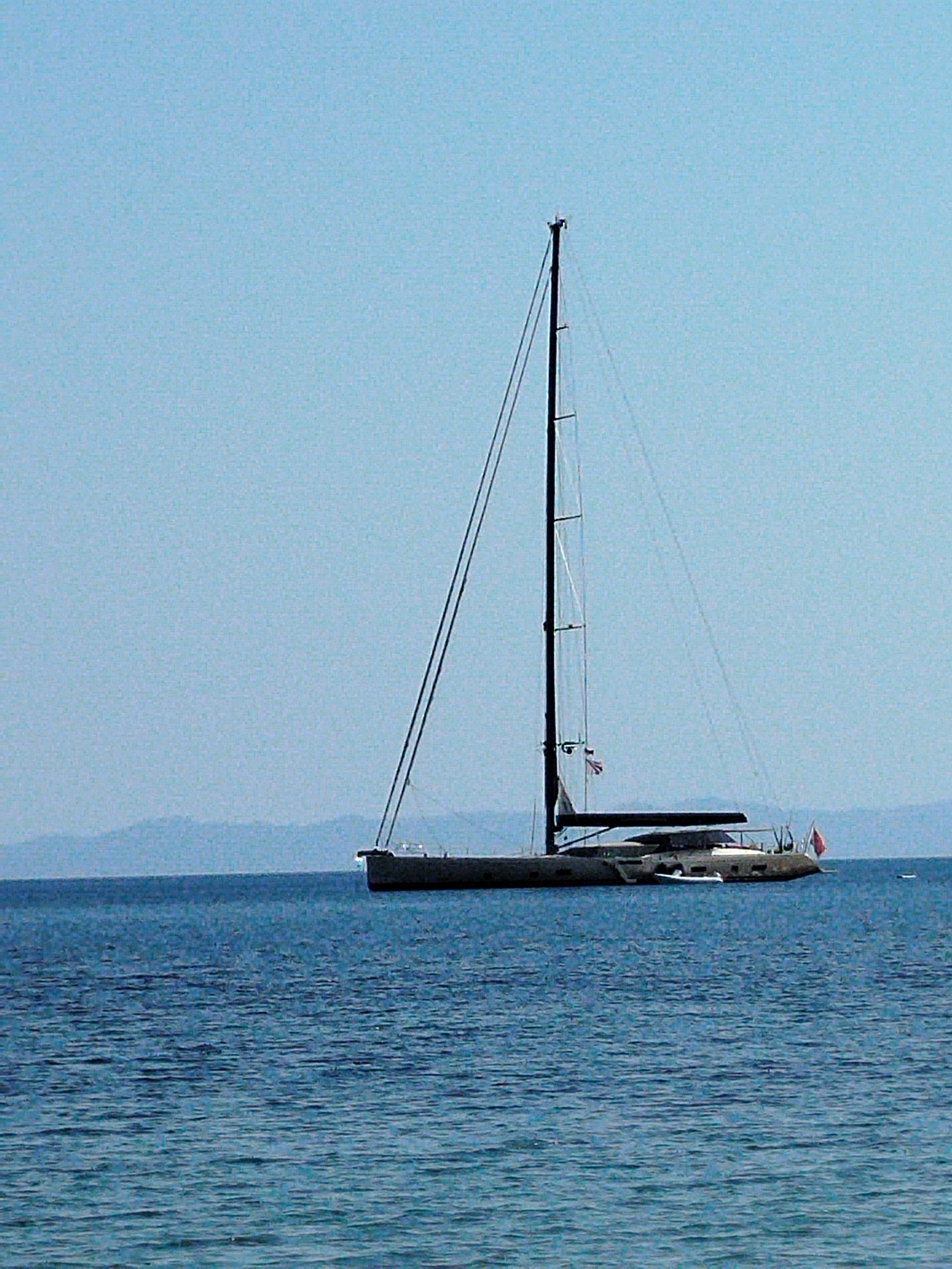 A wide shot shows a large sailboat anchored in calm, blue waters. The sailboat, with its dark hull and tall mast, is positioned in the center-right of the frame. A smaller white dinghy is attached to the side of the sailboat. In the background, a hazy, blue mountain range is visible on the horizon, suggesting a coastal location. The sky is a clear, light blue. The scene appears to be Sithonia Municipal Unit, Greece.