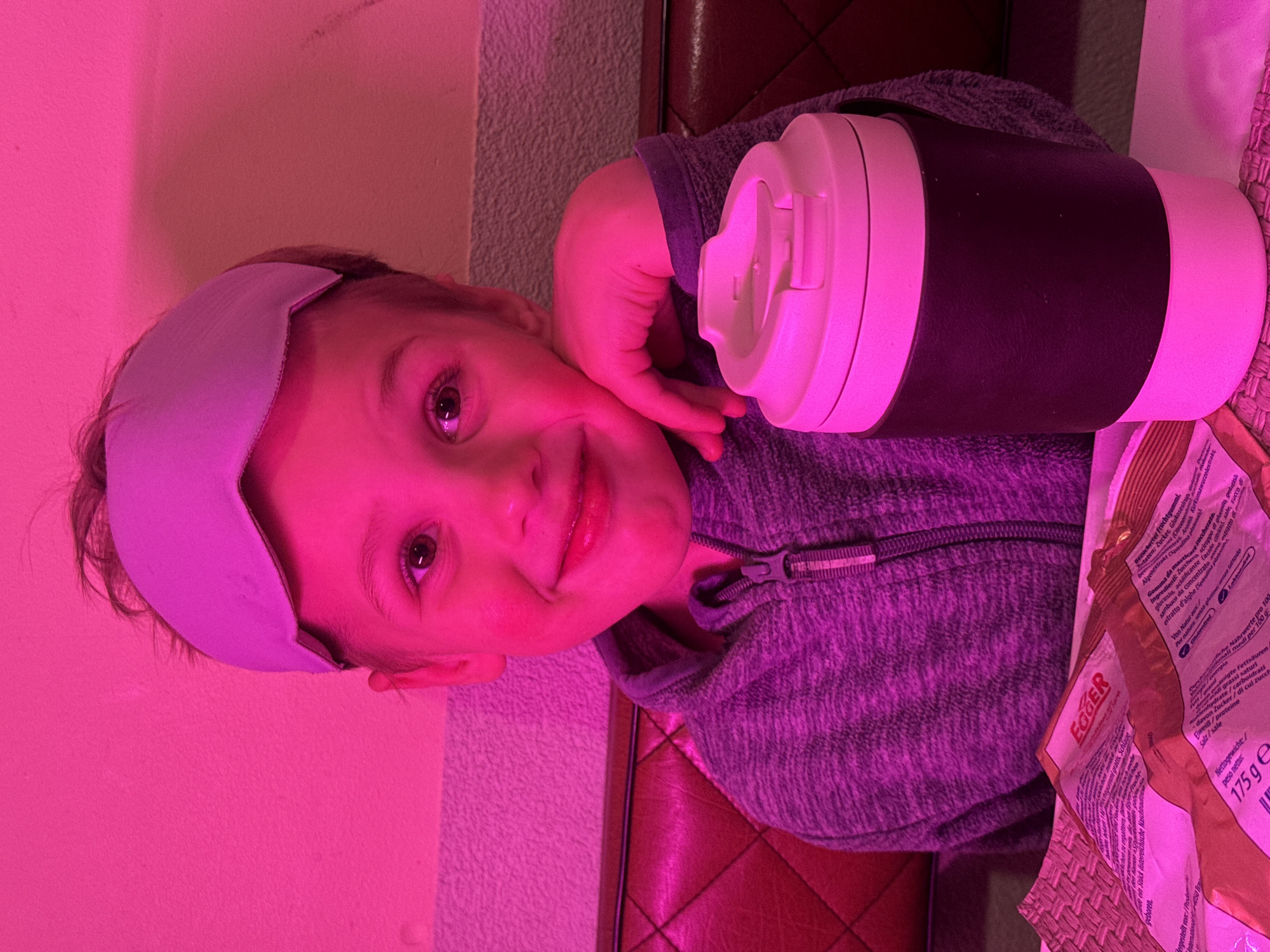A child, wearing a sleep mask, rests their head on their hand next to a black and white thermos and a package of Egger brand gum. The OCR data reveals text in German and Italian, suggesting the product is imported from Austria or Italy. The setting appears to be indoors, possibly on a couch or chair. The image is tinted with a pink or purple hue, which may be from the lighting or a filter. The location within Riga, Latvia, cannot be determined from the image.