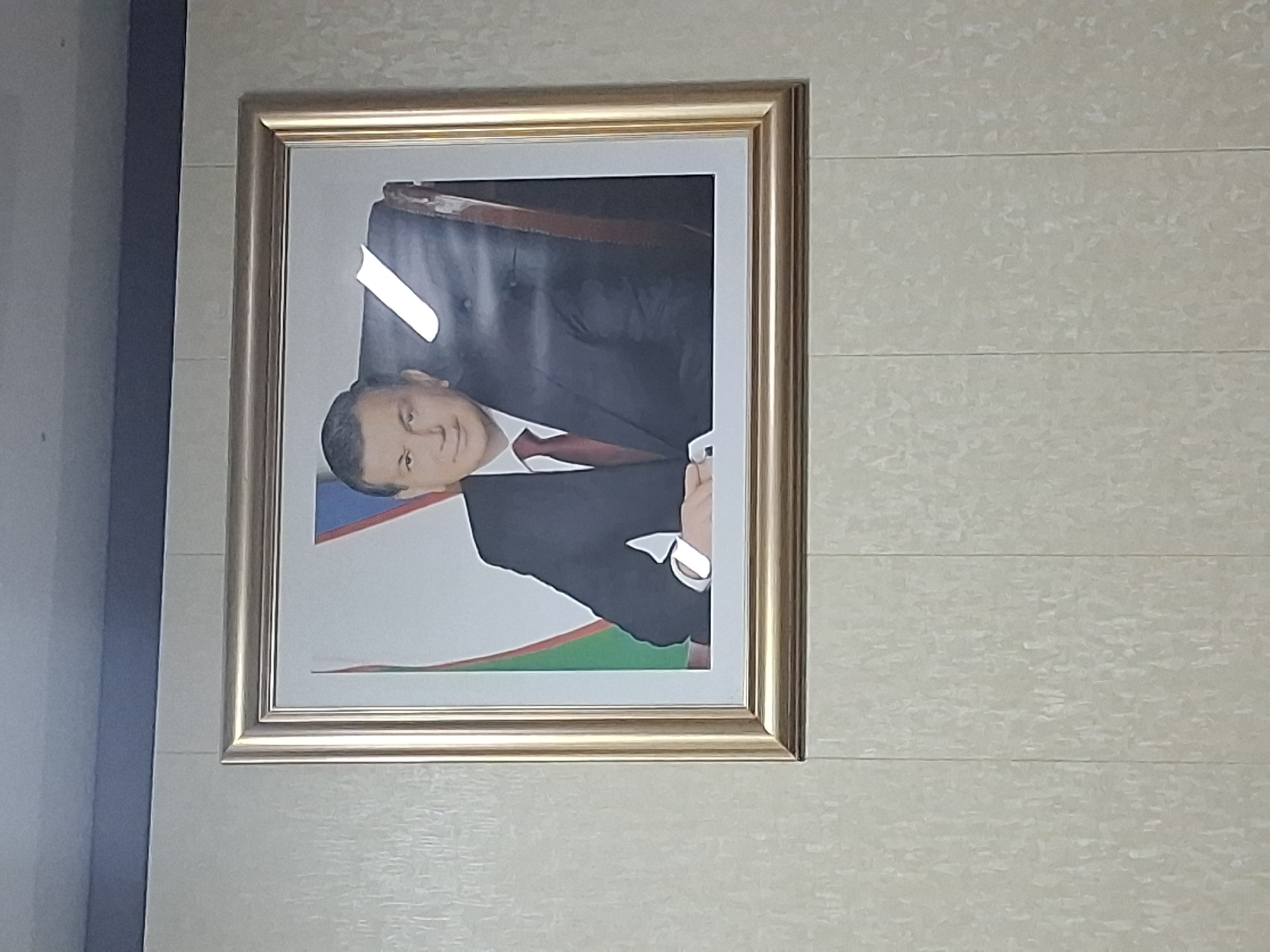 This media file shows a framed portrait of a man. The man is wearing a dark suit, a white dress shirt, and a red tie. He is looking directly at the camera with a serious expression. The portrait is hanging on a wall with a textured, light-colored wallpaper. The frame of the portrait is metallic gold. The background behind the man appears to be a flag, featuring blue, white, and green stripes with red crescent moons and stars. The overall lighting suggests an indoor setting, possibly an office or official building. The image is oriented vertically, and there is a bright reflection on the glass of the frame, obscuring a portion of the background. The provided location context is Kahramon, Uzbekistan, suggesting the portrait is of a prominent figure, likely a political leader or official, from that region.