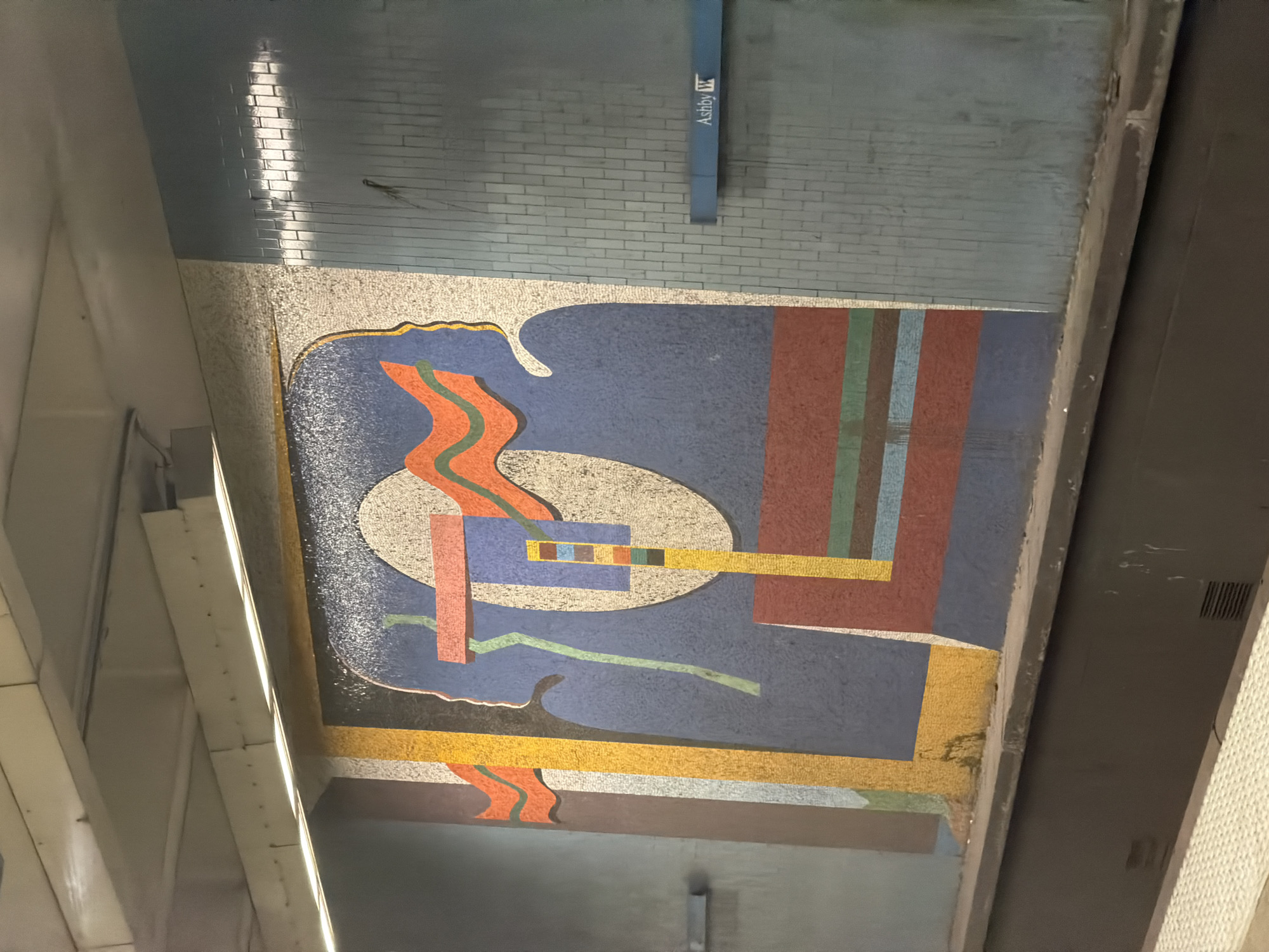 The image shows a mosaic art piece above the Ashby MARTA station in Atlanta, Georgia, USA. The mosaic consists of an abstract design with geometric shapes and wavy lines in orange, blue, yellow, and brown. It is situated above a platform or walkway, with structural support beams visible on either side. The surrounding infrastructure includes a brick wall painted in a light blue hue with a sign that says "Ashby W".