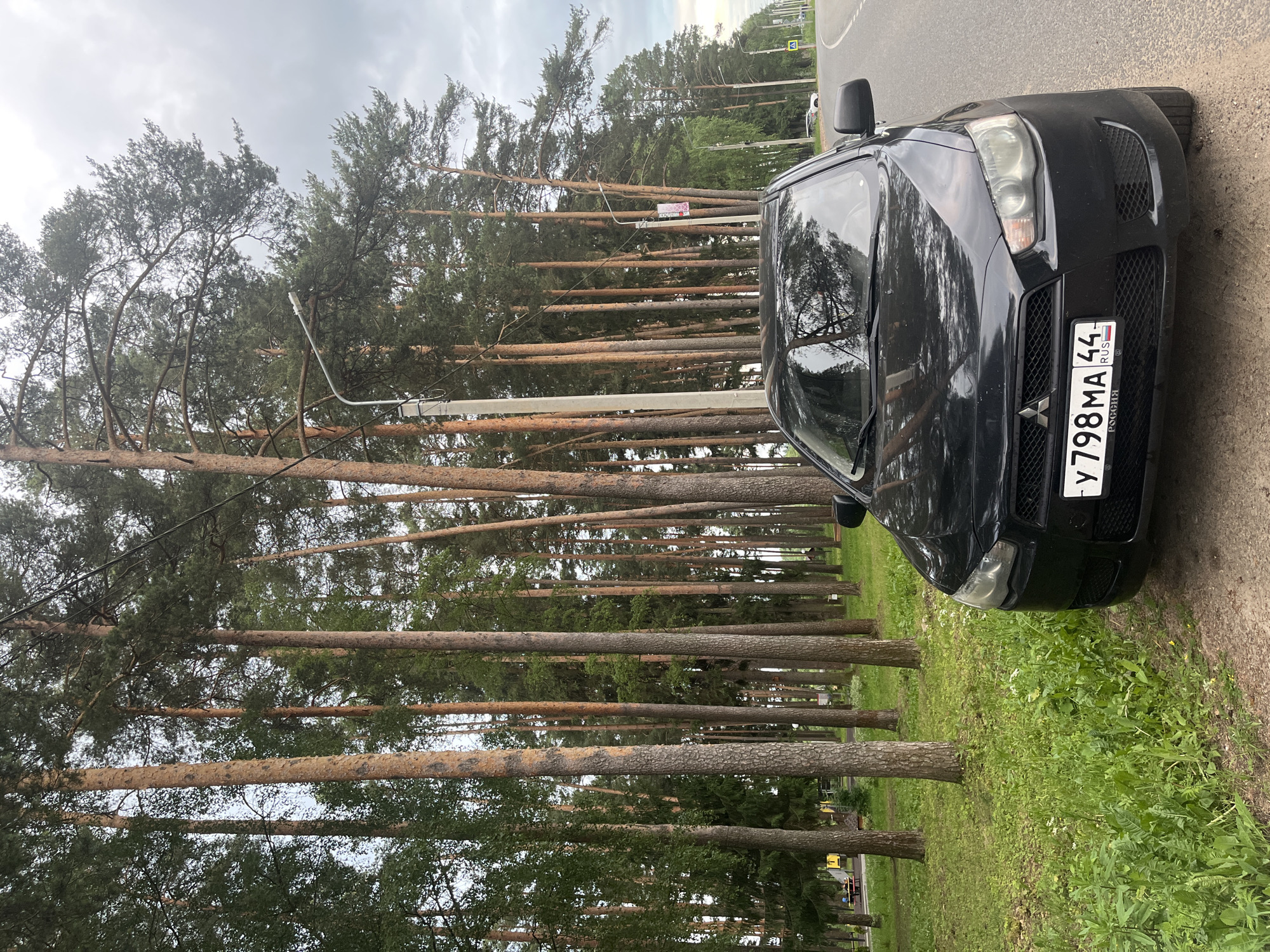 An eye-level, medium shot captures a black Mitsubishi Lancer parked on the side of a road, bordered by a dense forest of tall pine trees. The car is positioned at an angle, facing slightly left, with its front bumper and license plate clearly visible. The license plate reads "Y 798 MA 44" and has "РОССИЯ" written below it, indicating its Russian origin. The road to the right of the car is asphalt and curves away into the distance. In the background, to the left of the trees, a children's playground with yellow structures and red picnic tables can be seen. The sky is overcast with grey clouds. The overall scene is in the city of Nikolskoye, Russia.