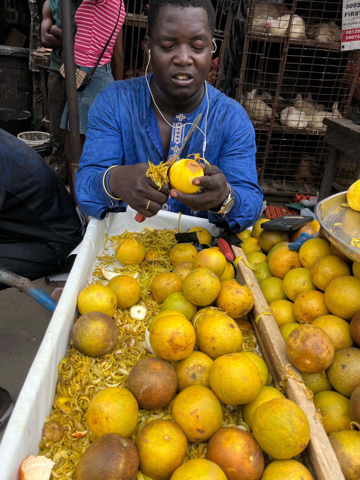 A Nigerian man in a blue tunic with intricate white embroidery and a gold bracelet is peeling an orange in the foreground. He wears white earbuds. He is in a market, likely in Ado, Nigeria, as evidenced by the presence of live chickens in cages behind him, a common sight in Nigerian markets. The man is surrounded by a large display of oranges, some of which have been peeled and their zest discarded. In the background, other market-goers are visible, including a person in a pink and white striped shirt. The overall scene depicts a vibrant market atmosphere.
