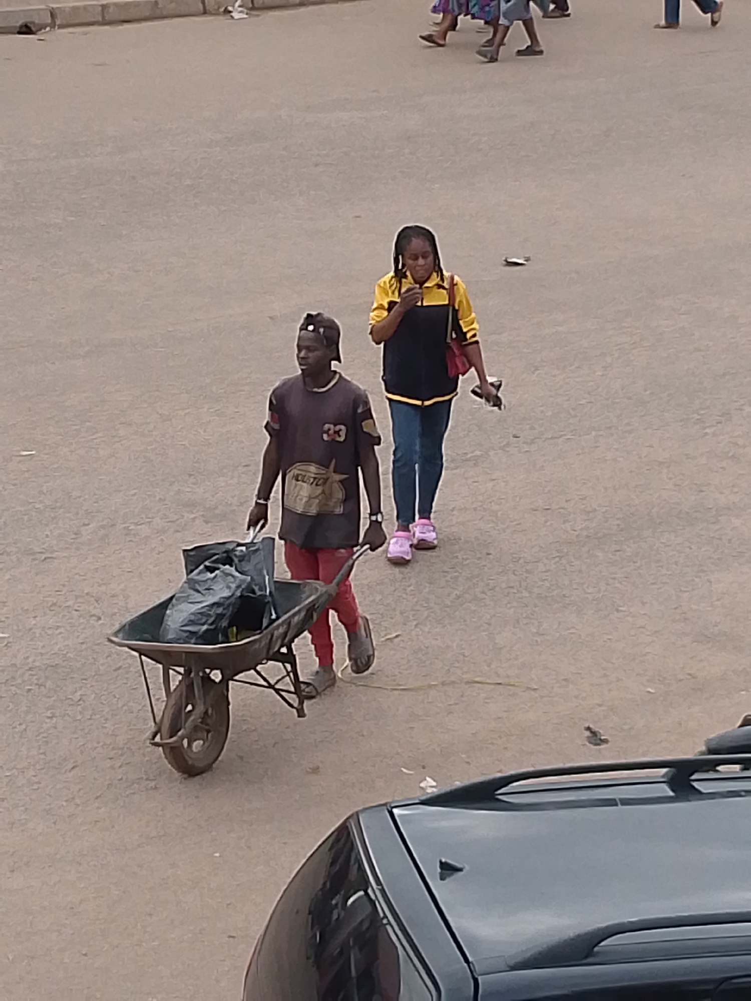The image depicts an overhead view of a street scene in Bukuru, Nigeria. A dark-skinned man, wearing a dark cap reversed, a dark t-shirt with a "33" and "HOUSTON" graphic, red pants, and grey sandals, pushes a dark metal wheelbarrow containing a large black plastic bag. He is positioned in the lower-middle left, moving towards the right. Behind and to his right, a dark-skinned woman with braided hair, wearing a yellow and black long-sleeved top over a dark garment, blue jeans, and pink athletic shoes, walks in the same direction. She carries a red shoulder bag and holds a dark object, possibly a phone, in her left hand while looking downwards. The ground is a light grey-brown, unpaved or worn asphalt surface with minimal debris. The upper portion of a dark-colored vehicle with roof rails is visible in the bottom right corner of the frame. In the distant background, several blurred figures and legs of pedestrians are discernible, with a light-colored concrete curb along the top edge of the road.