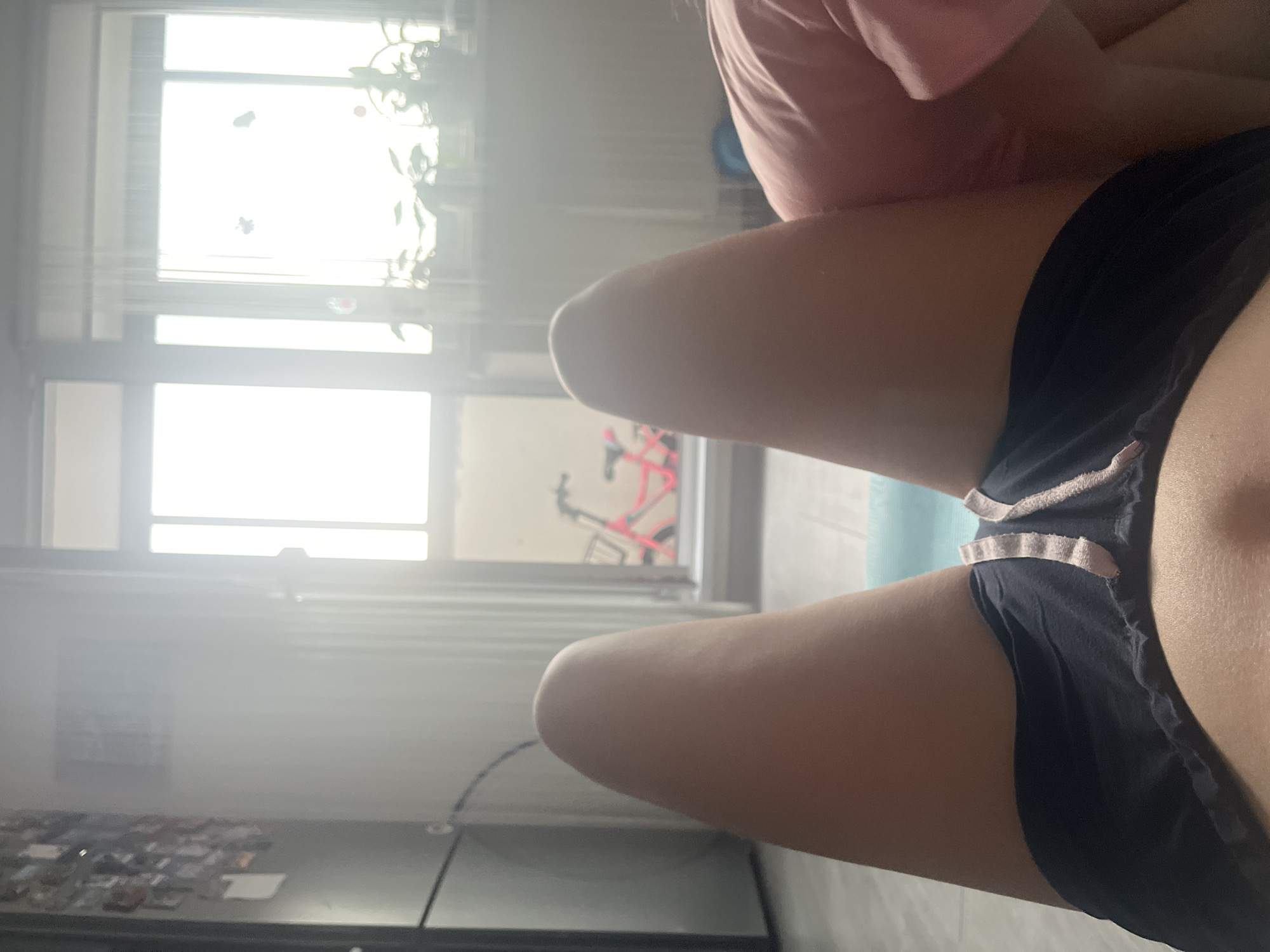 The media file is a close-up, angled shot of a person's legs and torso. The person is wearing dark shorts with light-colored trim. They appear to be sitting or lying down, with their legs bent and extended. The background is blurred but shows a window with blinds and some greenery, suggesting an indoor setting. Through the window, a bright light indicates it is daytime. A pink exercise bike is visible outside the window. The overall lighting is bright and somewhat overexposed, with a lens flare creating a hazy effect. No text is visible in the image. The location is Chelyabinsk, Russia.