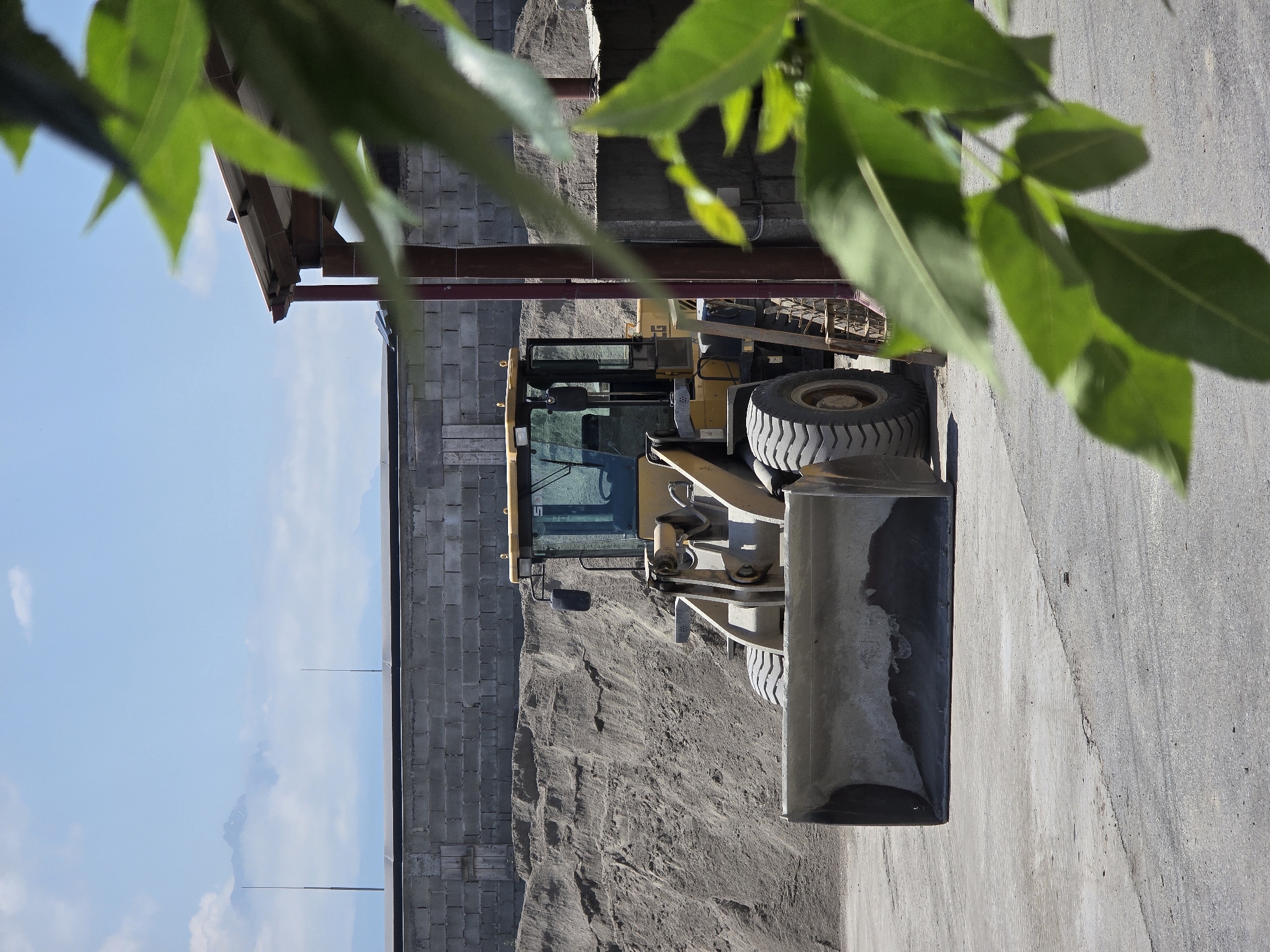 An outdoor, daytime scene shows a yellow wheel loader parked on a concrete surface in front of a large pile of sand and a concrete block wall. The wheel loader is facing forward, with its bucket lowered to the ground. To the right of the wheel loader, a building with a sloped roof supported by reddish-brown poles is partially visible. Out-of-focus green leaves from a foreground plant obscure a portion of the right side of the image. In the background, a low, gray concrete block wall runs horizontally, with mountains and a hazy sky visible beyond it. The scene appears to be a construction or material yard.