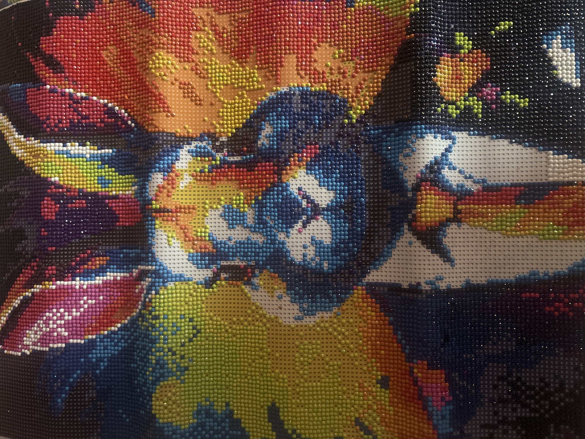 A colorful, partially completed diamond painting is depicted, showcasing a stylized or abstract image, possibly of an animal's face or a creature, with prominent features. The "eyes" are formed by blue and white beads, while the surrounding elements are rendered in vibrant reds, oranges, yellows, purples, and blues, set against a dark, almost black background.
The artwork is made up of countless small, shiny, round resin beads ("diamonds" or "drills") affixed to a canvas. Large sections are densely covered with these beads, creating a textured and sparkling surface that reflects light. Other areas of the canvas are still unfinished, revealing a printed grid with various symbols. These symbols guide the placement of specific colored beads.
No direct activity is taking place; the image is a static display of the artwork in progress. The notable details include the vivid, contrasting color palette, the shimmering quality of the beads, and the visible grid lines with symbols on the uncompleted portions. Visible symbols include 'H', 'G', 'L', 'J', 'X', 'V', 'C', '7', '6', '9', which indicate bead colors for the diamond painting pattern. The image does not contain any specific visual cues that link it to Ruse, Bulgaria.
