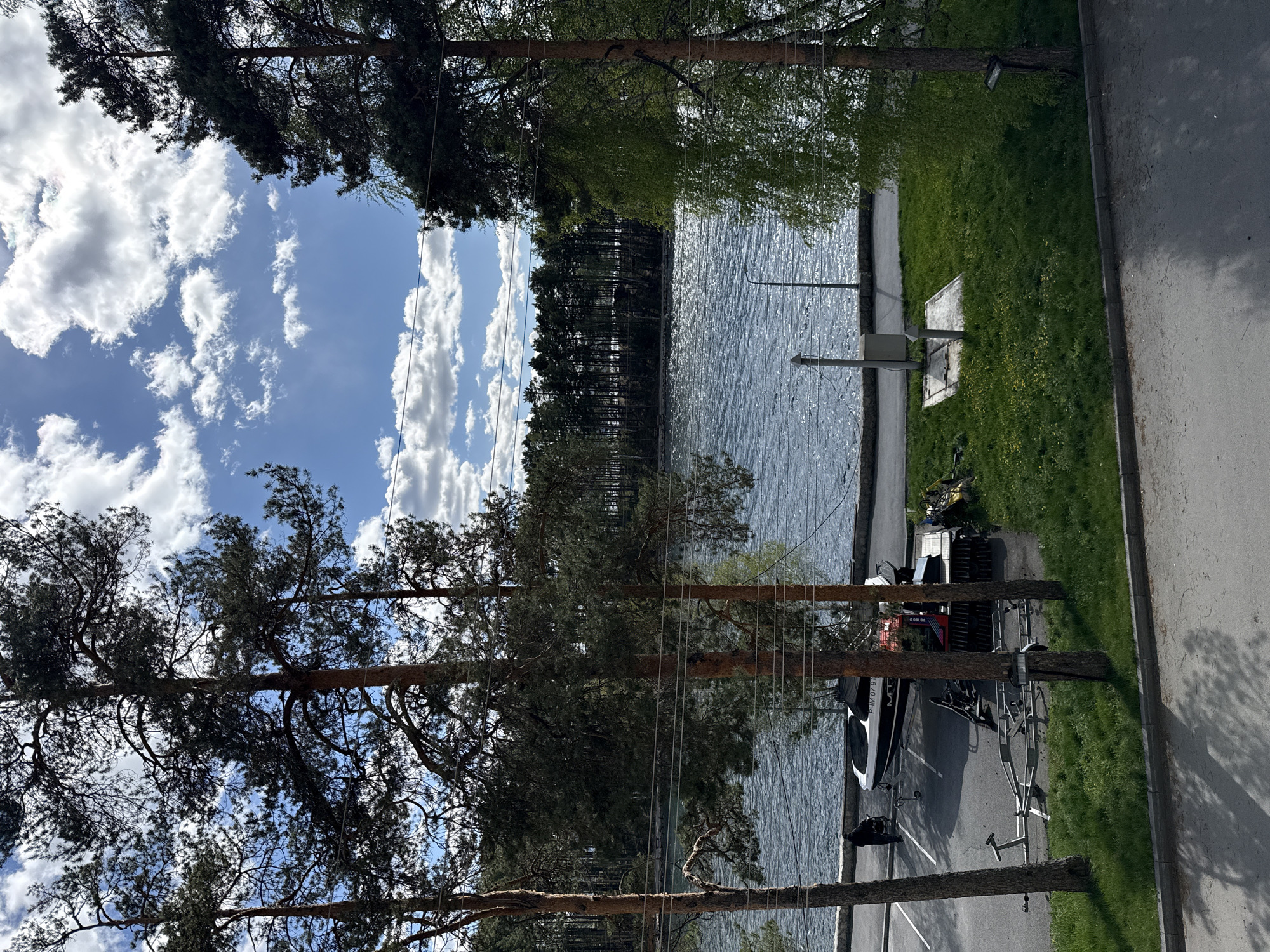 The image shows a waterfront scene in Turgoyak, Russia. A body of water, likely Lake Turgoyak, is visible with a treeline in the distance across the water. Closer to the viewpoint, there is a grassy area and a paved parking lot. In the parking lot, a boat on a trailer is parked, along with a vehicle. Trees are visible in the foreground, framing the scene. The sky is blue with scattered white clouds.