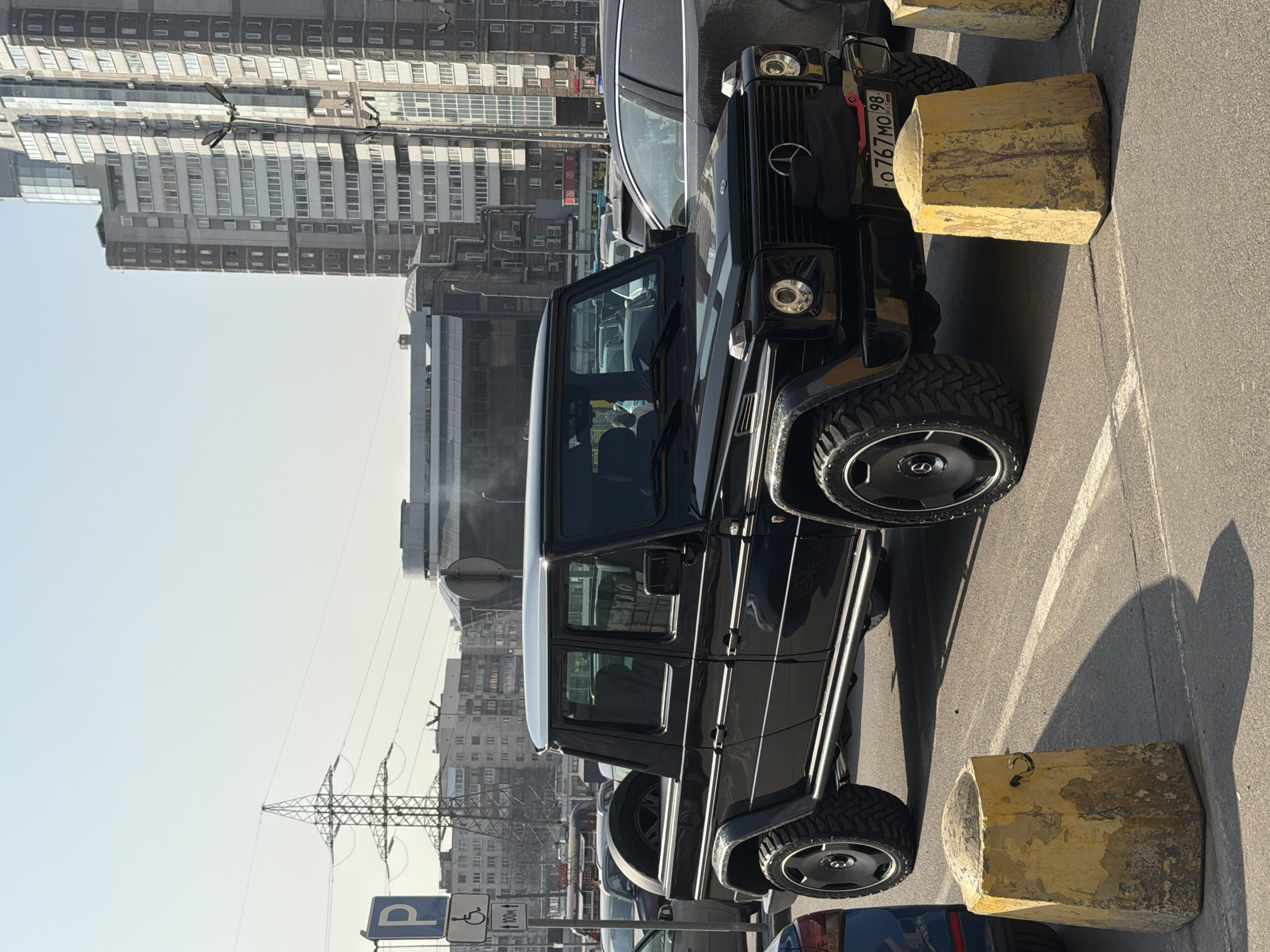 The image depicts a black Mercedes-Benz G-Class SUV parked between two yellow concrete bollards in Saint Petersburg, Russia. The license plate, though partially obstructed, appears to be Russian with the code "98." In the background, there are several multi-story buildings and an electrical tower. The sky is light blue, suggesting a clear day. The shadows are sharp, indicating a strong light source. A parking sign is visible in the lower left corner, including the disabled parking symbol. Other cars are parked nearby.