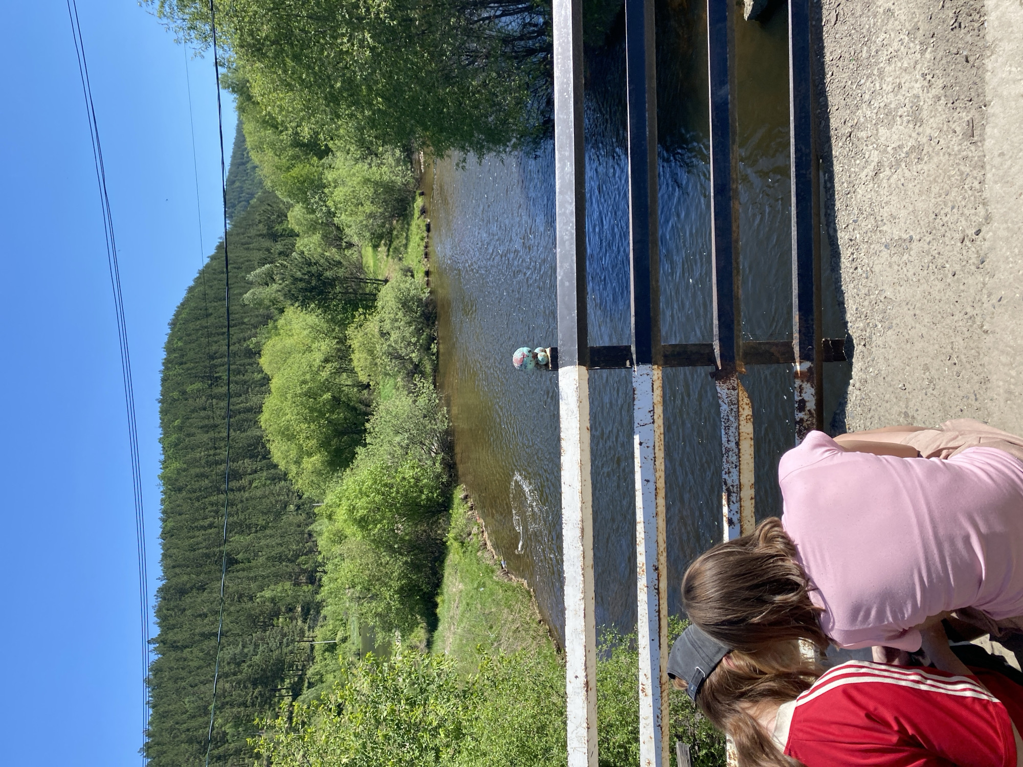 The image depicts a bright, sunny day on a bridge overlooking a river in the city of Bolshoy Lug, Russia. Two individuals, viewed from behind, are seated on the bridge, looking towards the water. One person wears a red jacket with white stripes, and the other wears a pink t-shirt. A small, globe-shaped ornament is visible on the railing of the bridge. The river flows through a lush, green landscape with dense forests covering the hills in the background. Power lines are strung across the sky. The bridge has a white railing at the top, with black metal bars below. The ground on the bridge is made of concrete.