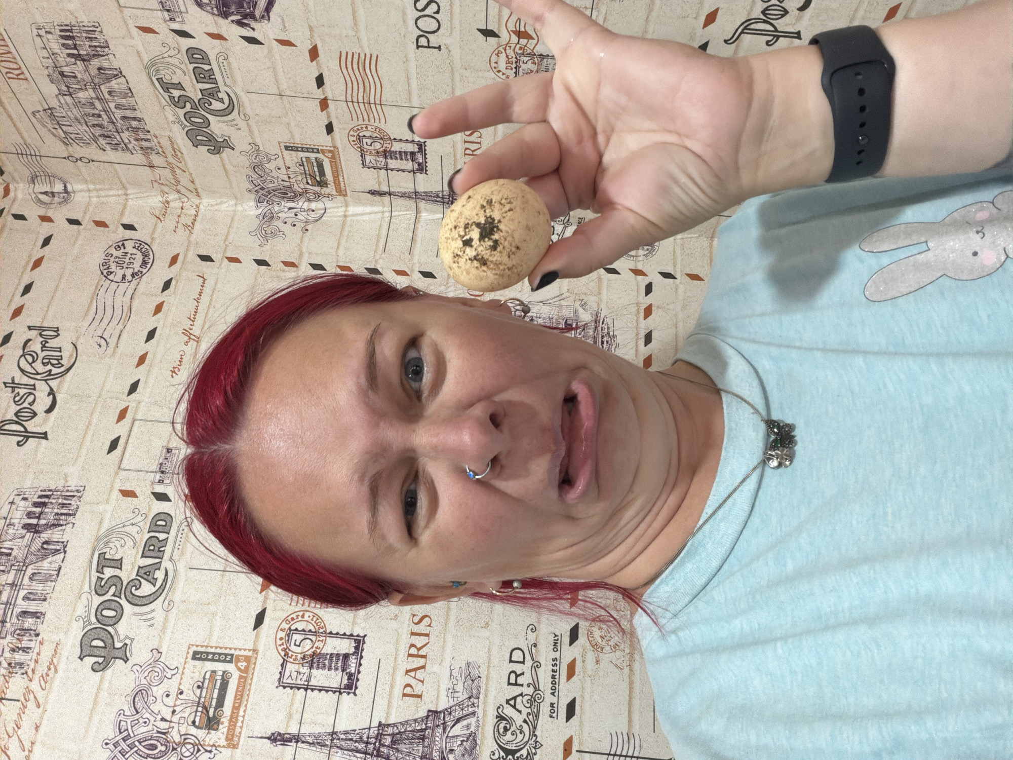 A woman with bright red hair and a grimacing expression holds up a speckled egg in her right hand. She is wearing a light blue t-shirt adorned with a cartoon bunny graphic and a silver necklace featuring two butterfly charms. The background is wallpaper depicting vintage postcards with images of famous landmarks like the Eiffel Tower and the Colosseum, along with postage stamps and the words "Post Card." The image appears to be a selfie, with the woman looking directly at the camera. There is no discernible location in the city of Chaikovsky, Russia.