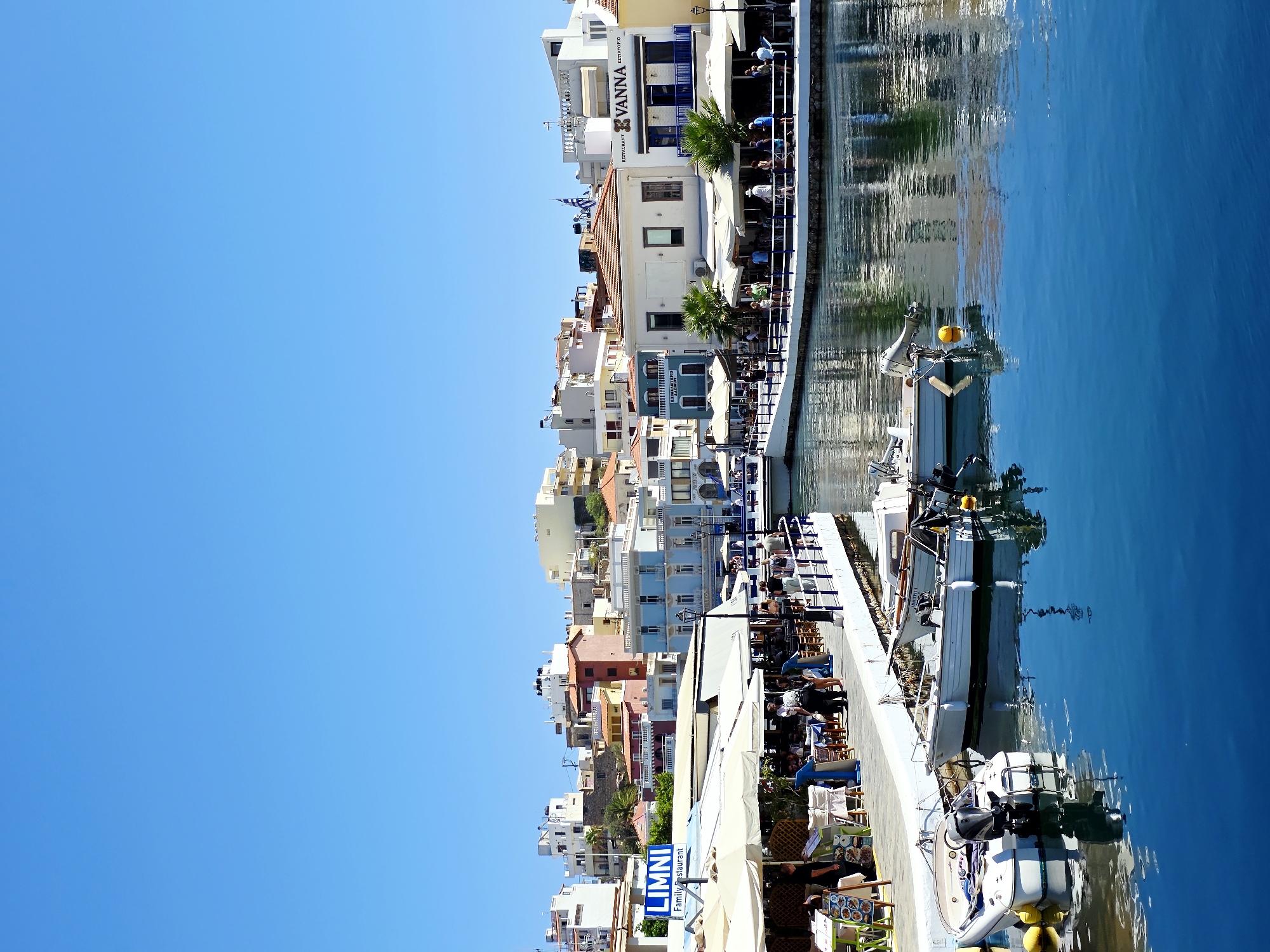 The image depicts a bright, sunny day in the city of Agios Nikolaos, Greece. The scene is centered around a picturesque harbor filled with clear, blue water. Several small boats with outboard motors are moored along a white dock. The dock is lined with restaurants and cafes, many of which have outdoor seating under white awnings. People are seen dining and strolling along the waterfront promenade. In the background, a colorful array of buildings, typical of Greek architecture, ascend a gentle slope. The sky is a vibrant, cloudless blue. The overall impression is one of a lively and inviting coastal town.