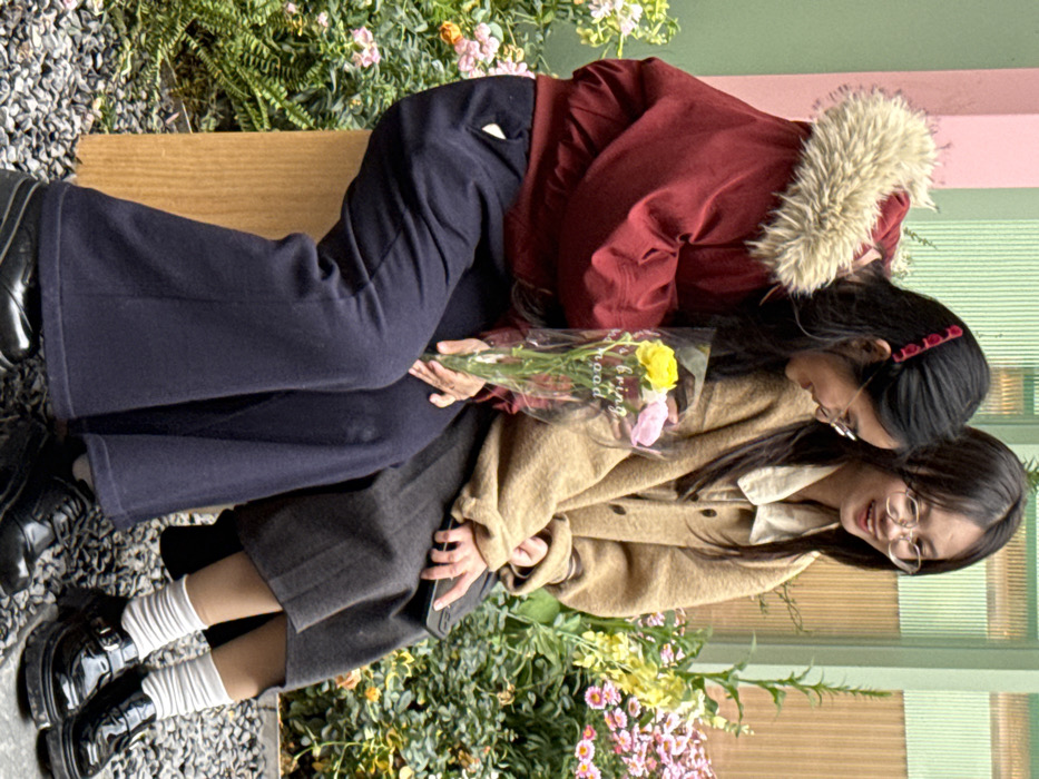 Two young East Asian females are seated on a light brown wooden block in what appears to be an outdoor environment in Liangjiang, China. The female on the left is wearing a dark red bomber jacket with a light brown faux fur collar, dark blue wide-leg trousers, and black platform lace-up shoes. Her dark hair features red decorative clips. She holds a small bouquet of yellow, pink, and white flowers encased in clear plastic, which has "bring mood" printed on it. She wears eyeglasses and is smiling, directing her gaze towards the female on her right. The female on the right is wearing a light brown button-up cardigan over a light-colored collared shirt, a dark grey midi skirt, white ruched socks, and black patent leather platform shoes with buckles. She also wears eyeglasses and is smiling, looking towards the female on her left. She holds a black smartphone in her left hand, resting it on her lap. The background consists of a light pink wall section to the left and a light green vertical ribbed wall section to the right. The immediate surroundings are filled with lush green foliage and various pink, yellow, and white blooming flowers. The ground beneath the subjects is covered with small grey pebbles.
