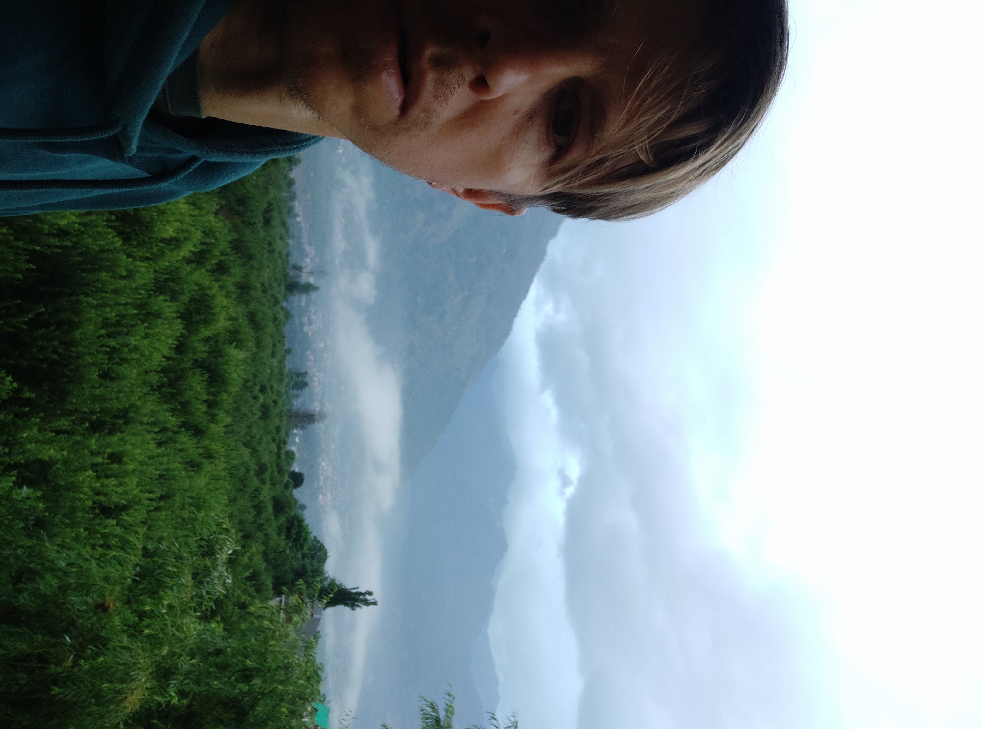 The image is a close-up, over-the-shoulder selfie taken in Patar, India. The left half of the frame shows the face and upper torso of a person wearing a teal hoodie. The person's face is turned towards the camera, with their left eye and part of their nose visible. Their hair is light brown and appears to be windswept. To the right of the person, the background reveals a lush green forest or orchard, which transitions into a valley with a small town or village nestled below. Beyond the valley, majestic, mist-covered mountains rise, their peaks obscured by heavy, grey clouds. The overall atmosphere is serene and slightly overcast, suggesting a cool or damp climate. The specific location is Patar, India.