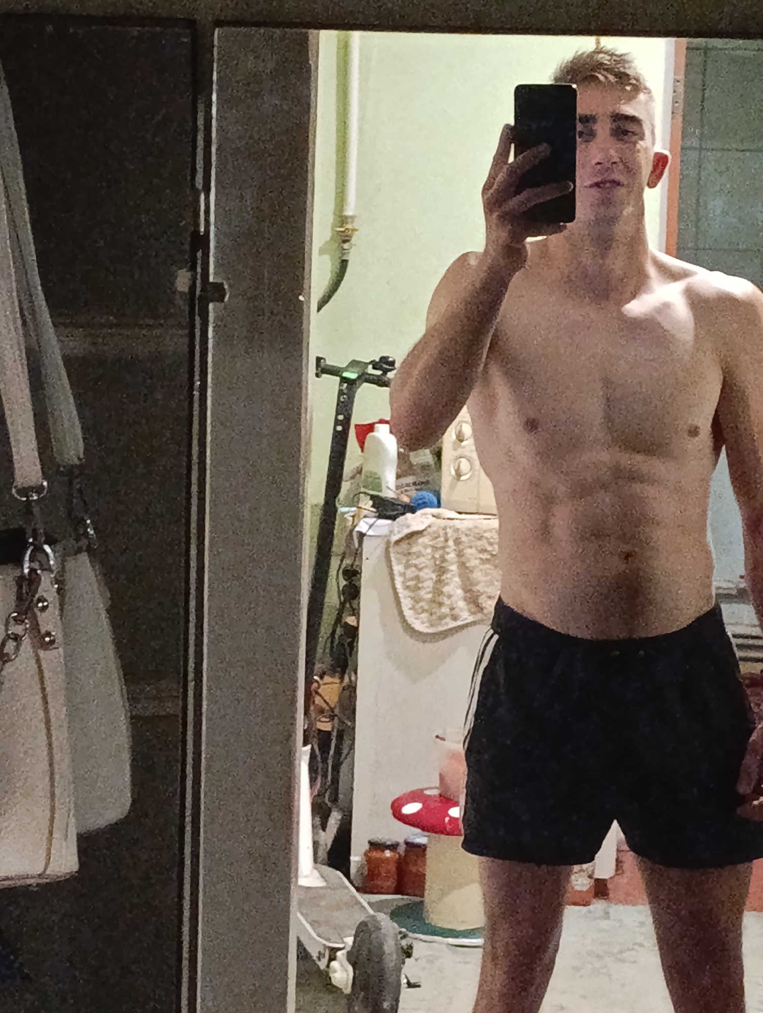 The image is a low-resolution, grainy selfie taken in what appears to be a residential setting, possibly a bathroom or dressing area. A young man with short, light-colored hair is visible from the chest up, holding a black smartphone to take the photo. He is shirtless, exposing a muscular torso, and wearing dark athletic shorts with white stripes on the sides. His expression is neutral, and he is looking towards the phone's screen.
In the background, a mirror reflects the man and the surroundings. The environment appears cluttered, with various objects visible on shelves and surfaces. These include cleaning supplies, a towel or cloth, and what looks like an electric scooter in the lower right portion of the frame. The lighting is dim, contributing to the grainy quality of the image.
While the image captures a personal moment, there is no specific information within the visual content that allows for an identification of the location within the city of Fastiv, Ukraine. The scene is generic and lacks any identifiable landmarks or characteristic features of that specific city.
