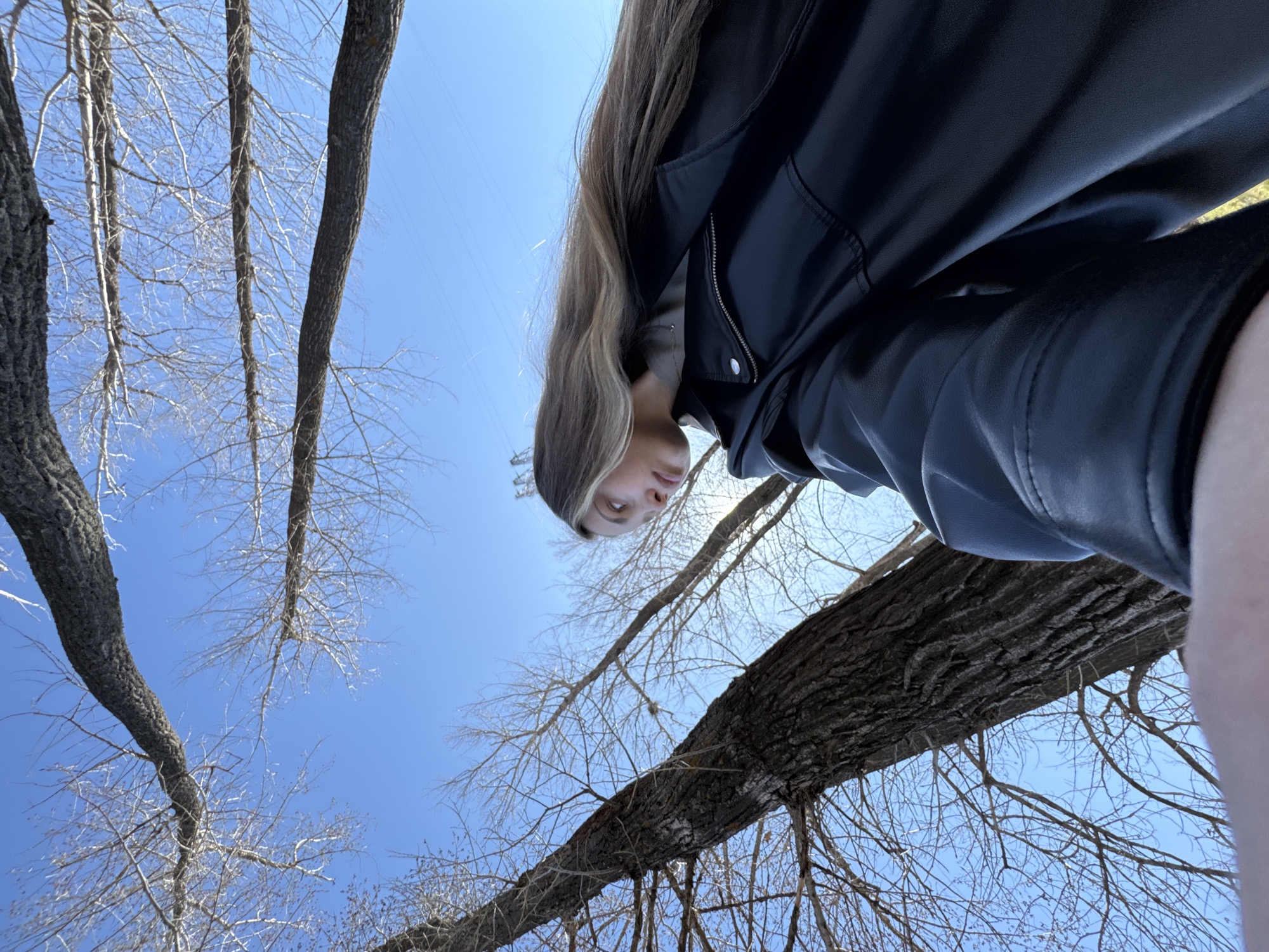 A low-angle shot depicts a young woman wearing a black leather jacket and a neutral-colored shirt in Chaikovsky, Russia. Her long hair is a blend of blonde and brown hues. She is positioned against a bright blue sky, which is partially obscured by the bare branches of trees. The perspective is from below, looking up at her. The surrounding environment appears to be a park or wooded area, as evidenced by the prevalence of trees.