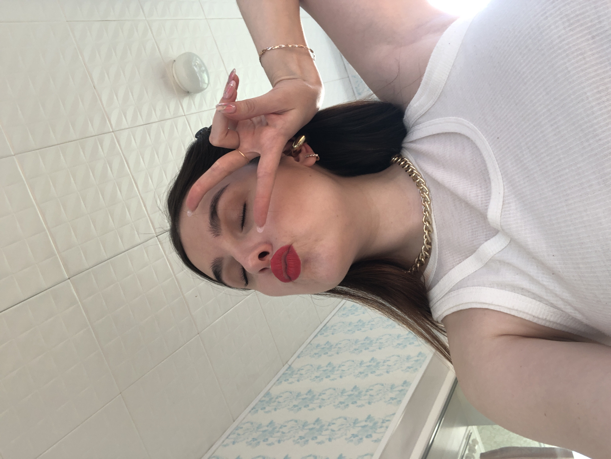 This image is a rotated portrait of a person, likely female, captured indoors in Noviy, Russia. The subject is shown from the chest up, featuring dark hair, fair skin, and vibrant red lipstick. She is dressed in a white ribbed t-shirt, accessorized with a prominent gold chain necklace around her neck, a gold chain bracelet on her right wrist, and multiple gold earrings or ear cuffs on her right ear. Her fingernails are long and manicured with a light-colored polish, possibly adorned with glitter or a subtle design.
The person is making a playful "peace sign" or "victory sign" gesture with her left hand, which is held up near her face. Her eyes are closed, and her lips are deliberately pursed in an expression often referred to as a "kiss face" or "duck face," conveying a relaxed, confident, or whimsical mood.
The background indicates an indoor setting, likely a bathroom, characterized by white tiled walls. The upper wall features white square tiles with a textured, raised diamond pattern. A white, circular wall-mounted light fixture is visible on this tiled surface. Below, a portion of the wall has a decorative light blue border or patterned tile with a delicate floral or scrollwork design. The lighting is bright and even, suggesting it is daytime. There is no visible text in the image.