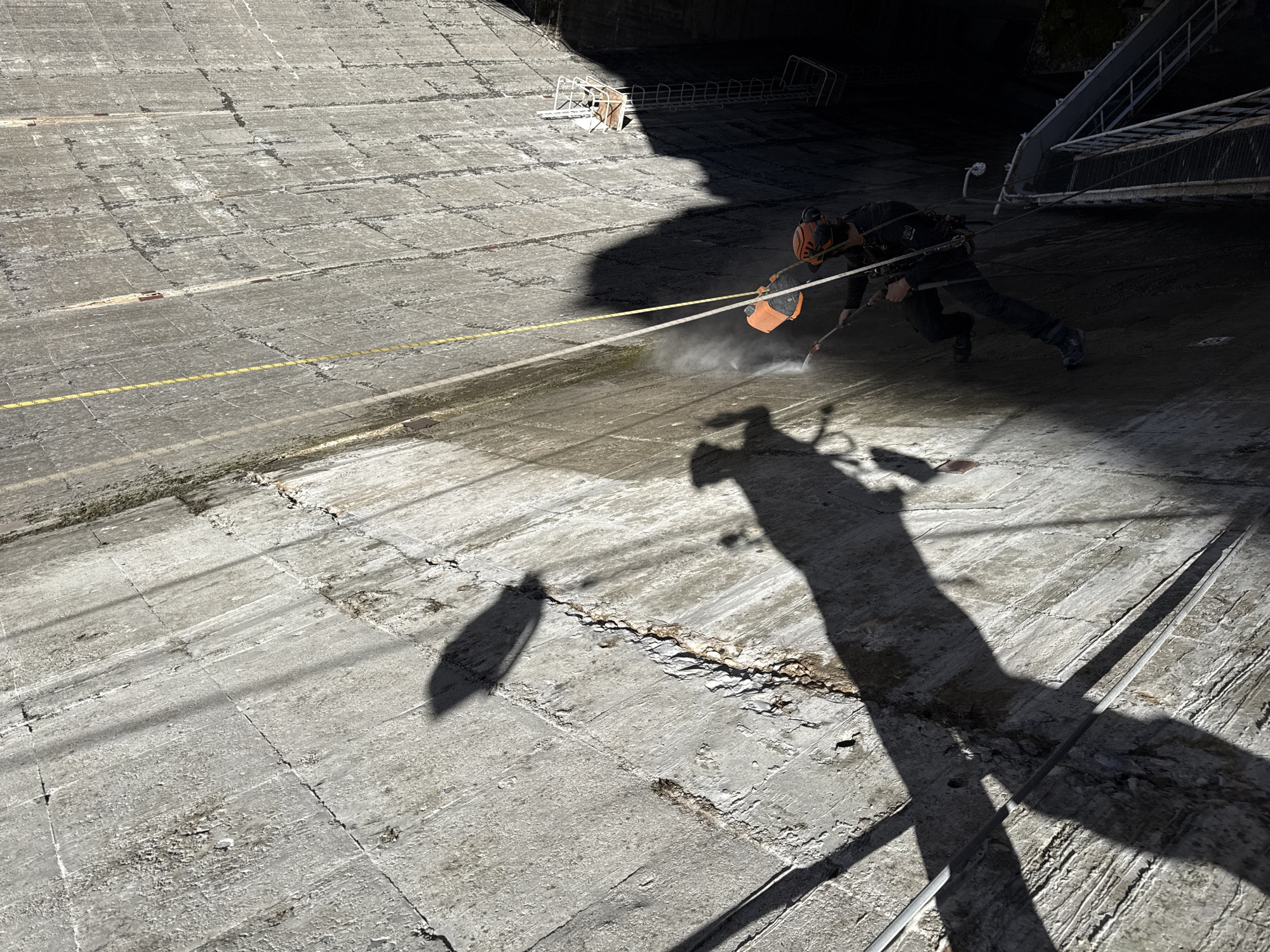 The image shows a person suspended by ropes, likely abseiling, cleaning a large concrete wall, possibly a dam. The person is wearing a helmet and safety gear, and appears to be using a water jet or similar device, as evidenced by the spray. The wall is made of large concrete blocks with visible seams and weathering. There are metal structures attached to the wall at the top right. The location is inferred to be Castellane, France, based on the information provided with the prompt and the visible concrete dam structure.